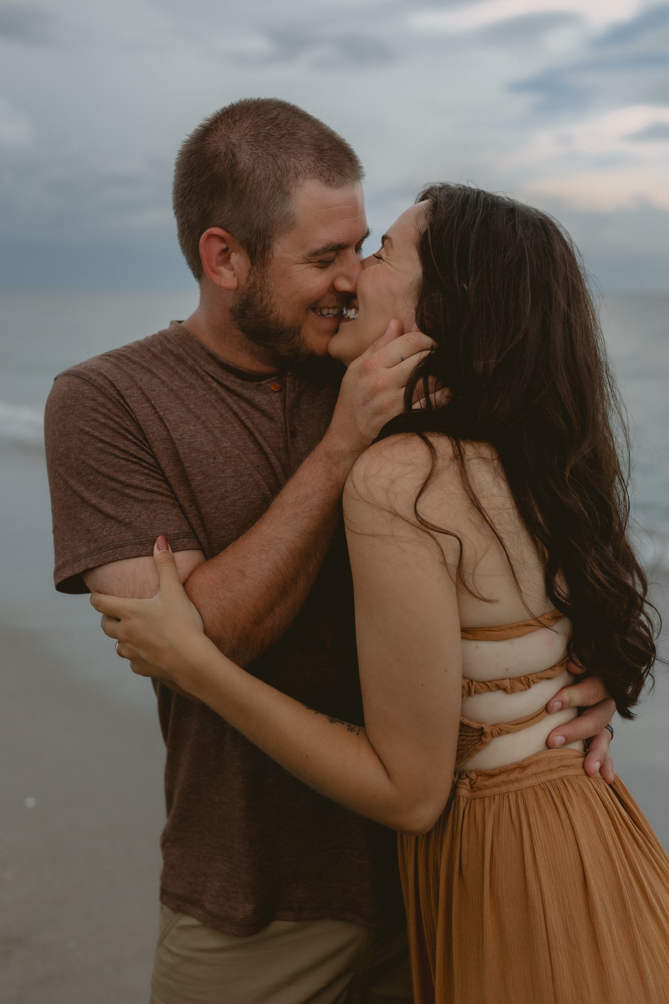 A couple kissing and embracing on the beach with ocean and cloudy sky in the background.