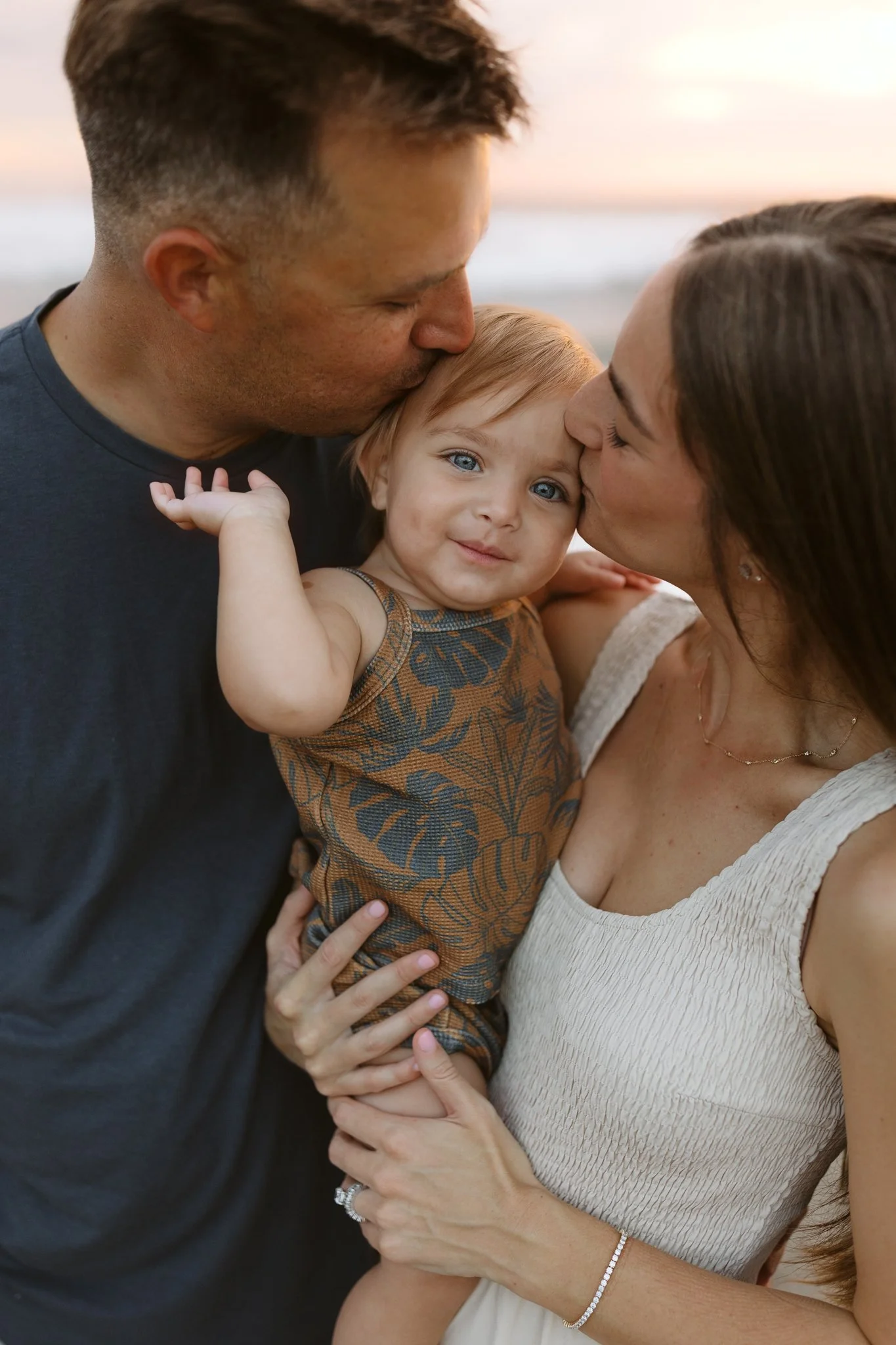 A family of three at the beach during sunset, with the father and mother kissing their young daughter on the cheeks.