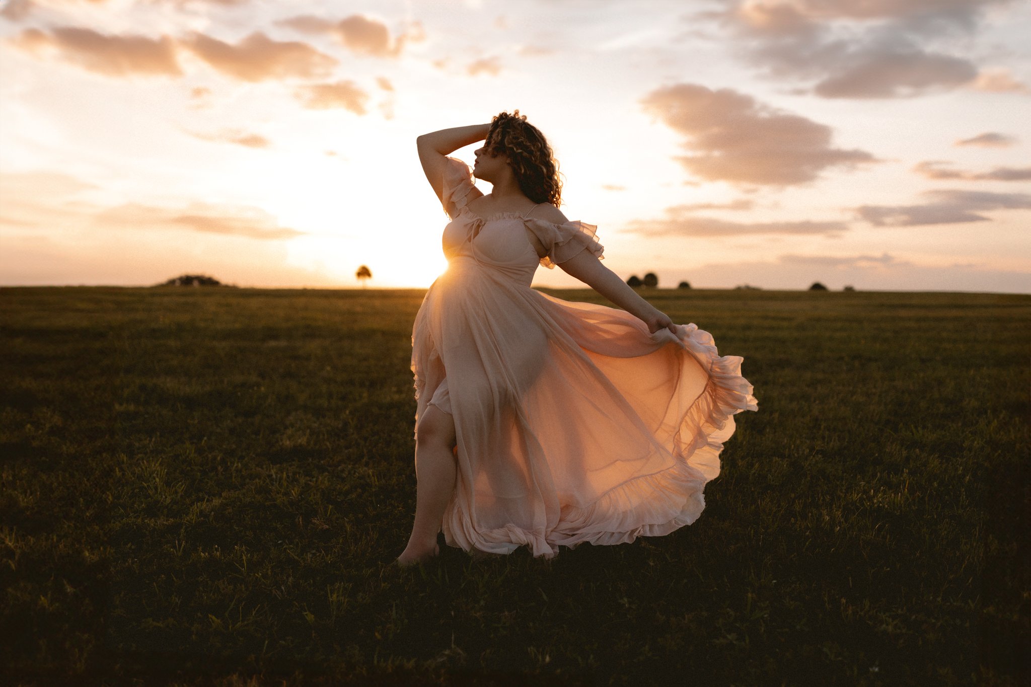 A woman wearing a flowing, peach-colored dress stands in a field at sunset, holding the skirt of her dress with one hand and her other hand resting on her head, with clouds and a few trees in the distance.