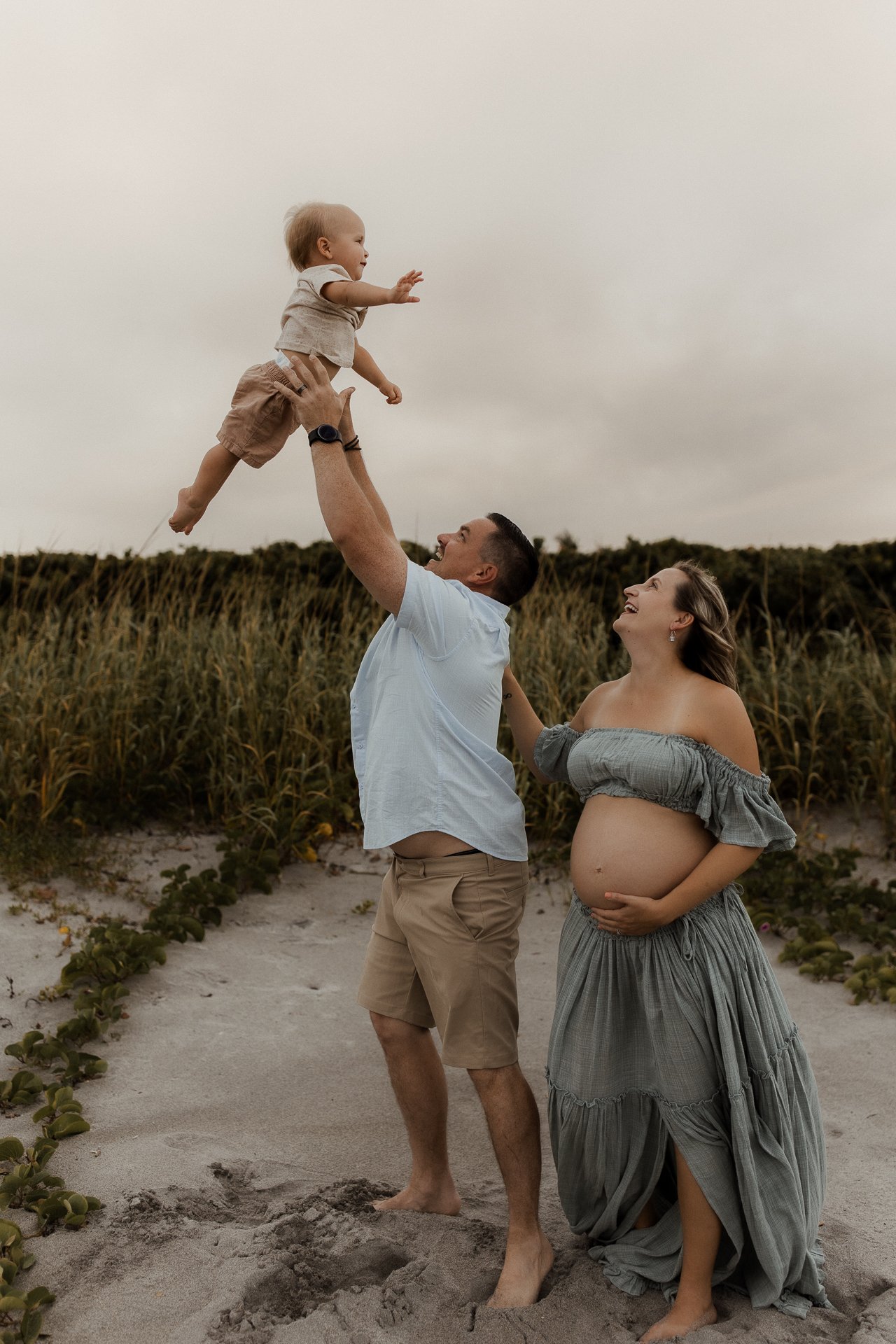 Family of three celebrating on the beach, with the father lifting the little child into the air and the pregnant mother smiling.
