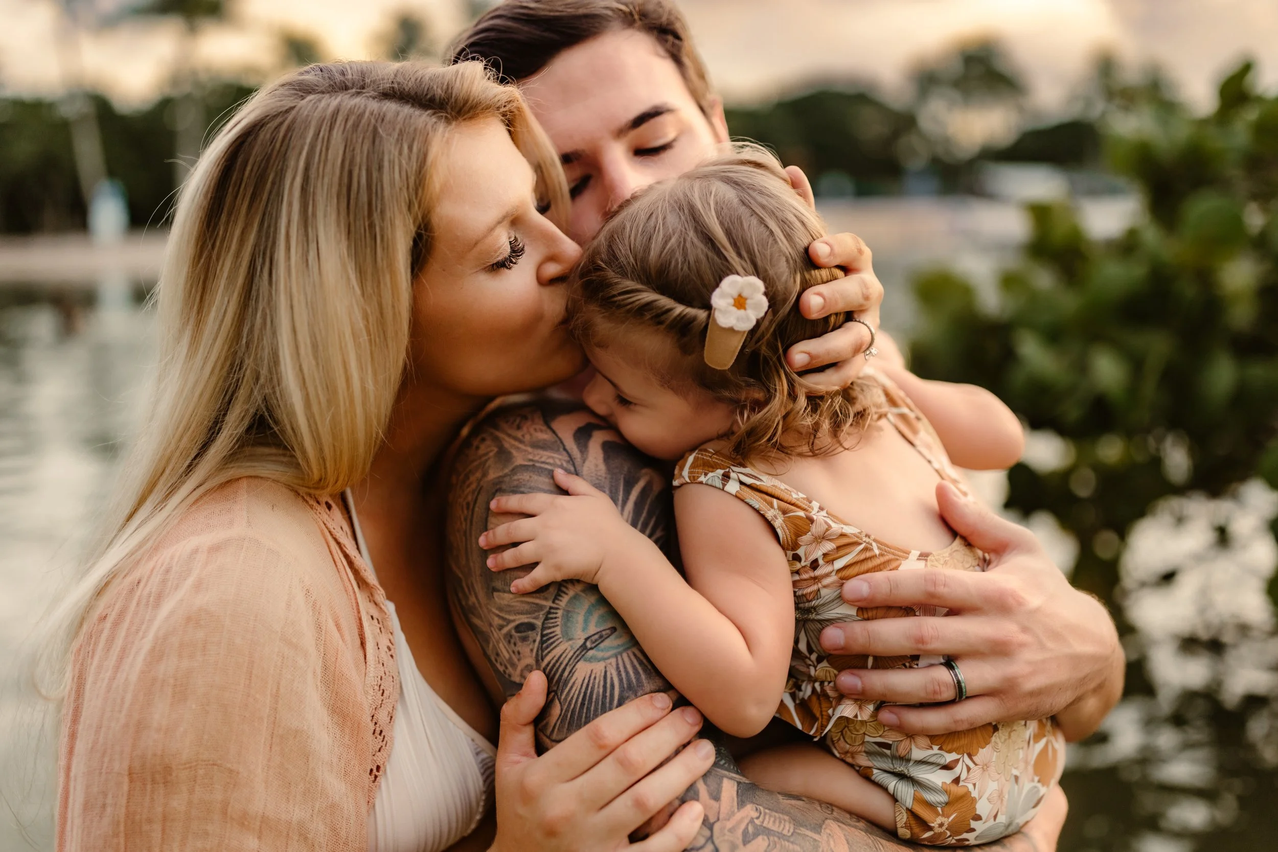 Four people sharing a group hug outdoors, one kissing a young girl on the forehead, two smiling and closing their eyes, with a water and greenery background.