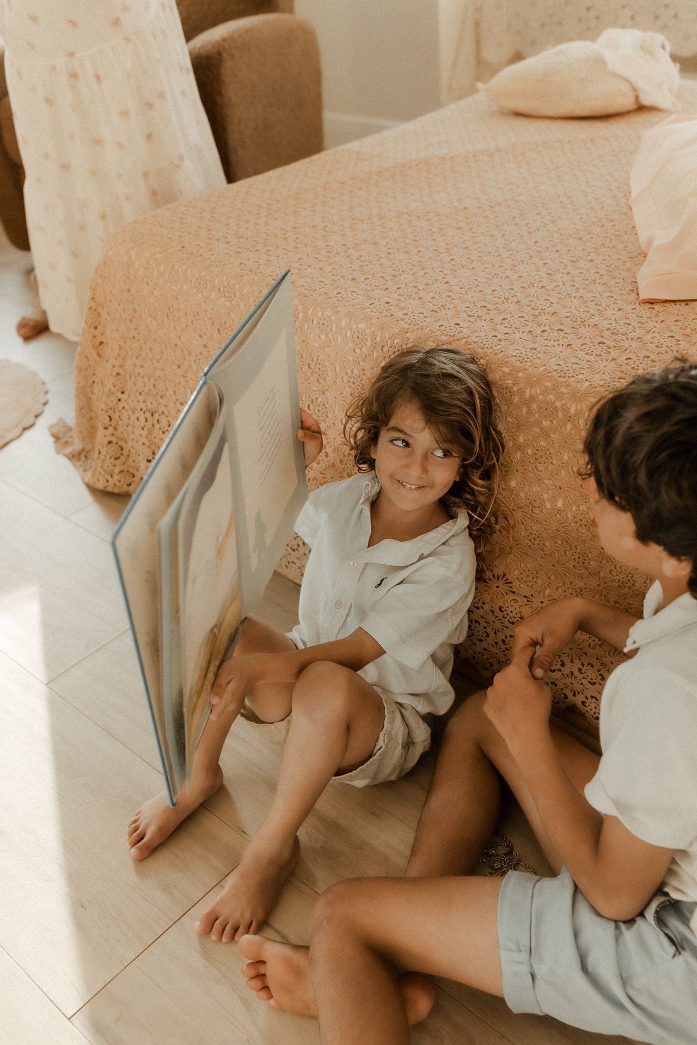 Two children, a girl and a boy, sitting on the floor next to a bed, engaging in a conversation. The girl is holding a large book, and they are both smiling.
