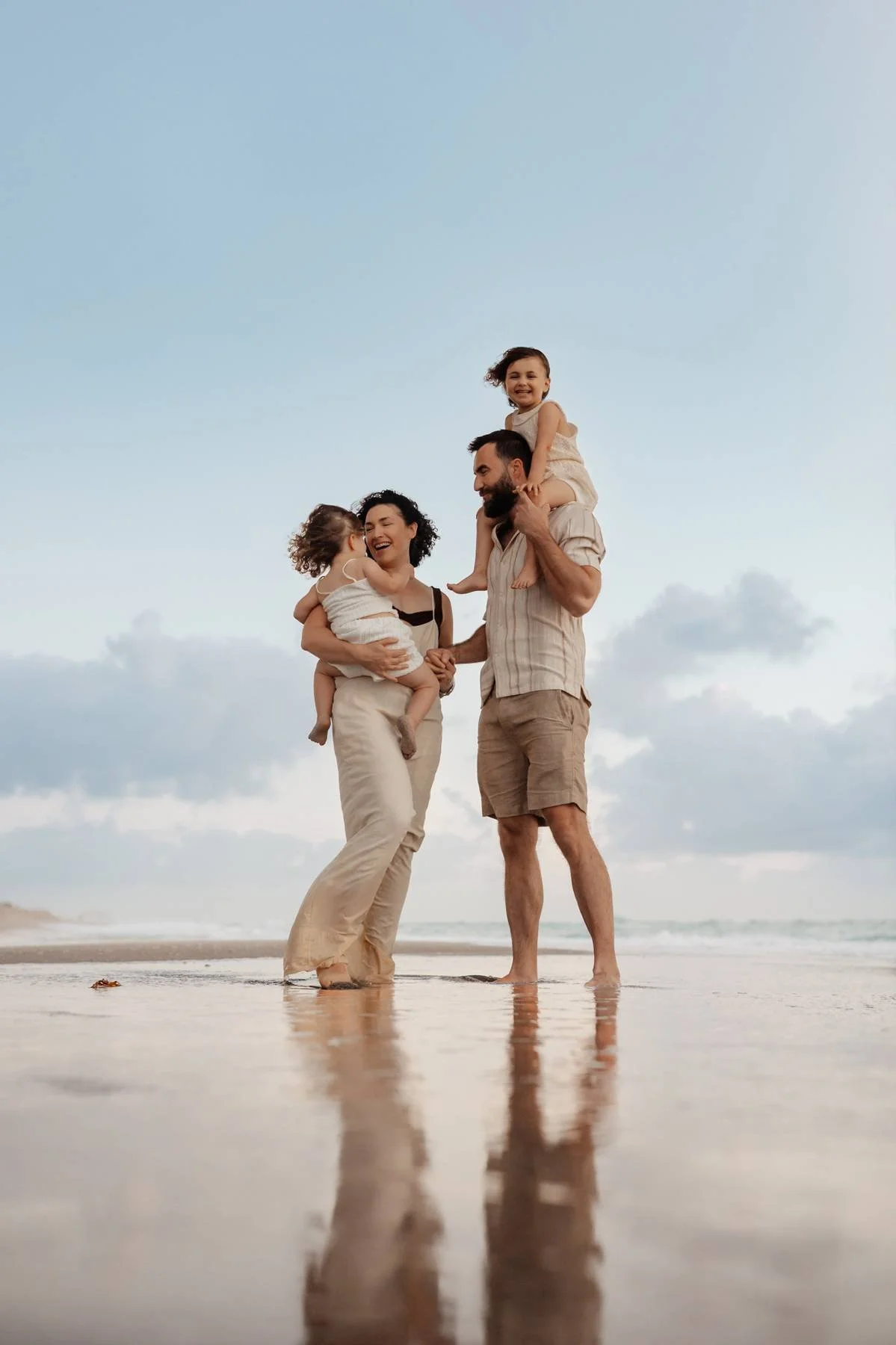 Family of four on the beach, two children on parents' shoulders, all smiling and enjoying the moment.