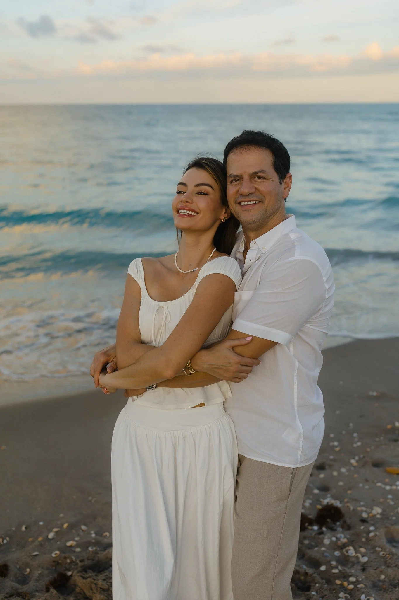 man bringing his wife in closely while smiling at the camera while standing on the beach