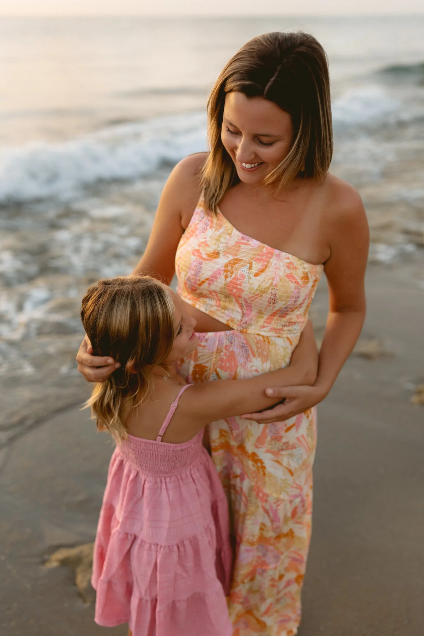 A woman and a young girl embracing on a beach at sunset, with ocean waves in the background.