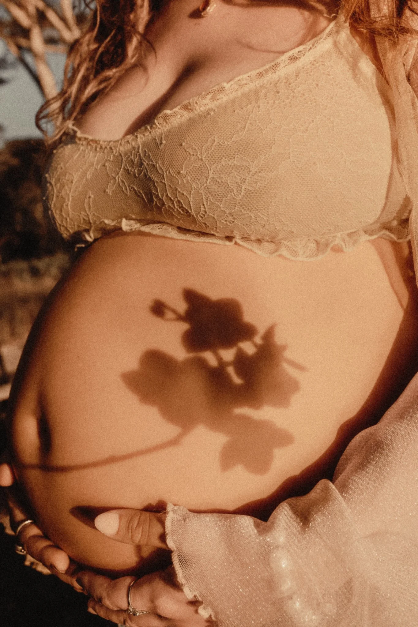 A pregnant woman wearing a lace top in warm lighting, holding her belly, with her hand showing rings, and the shadow of a flower on her stomach.