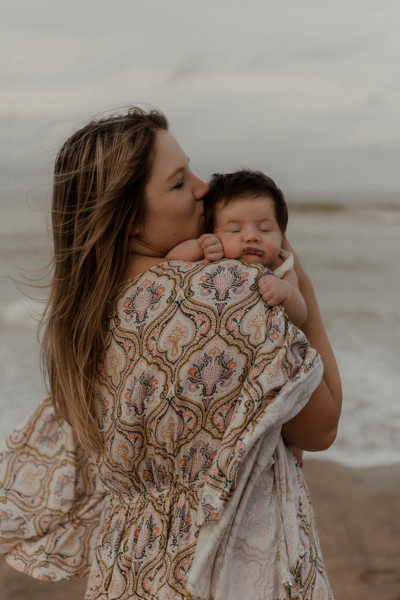 A woman holding a young child on a beach, with the woman kissing the child's head. The woman has long, brown hair and is wrapped in a patterned shawl, and the child has short dark hair and closed eyes.