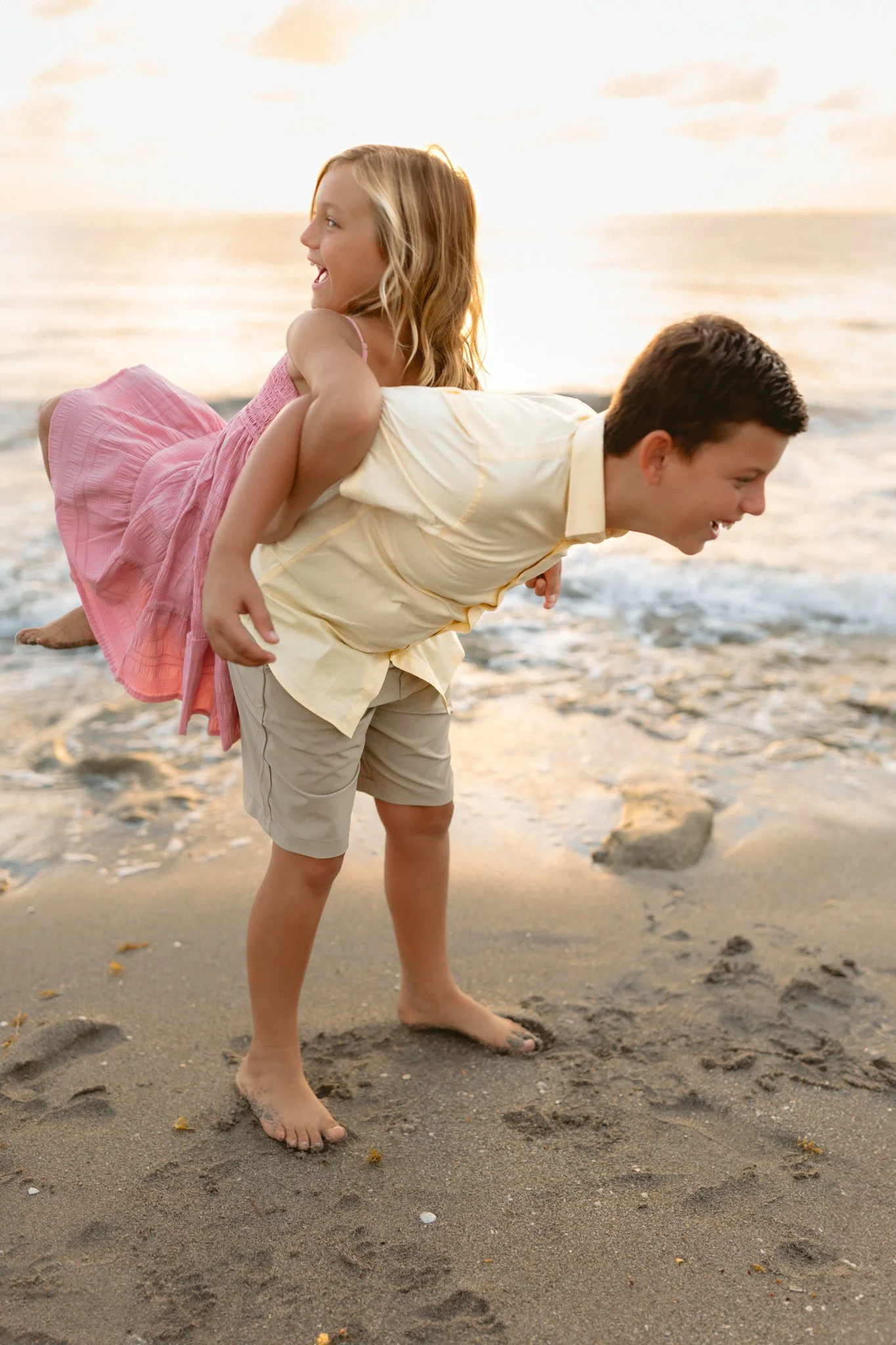 A young girl in a pink dress riding on the back of a boy in a yellow shirt and beige shorts at the beach during sunset.