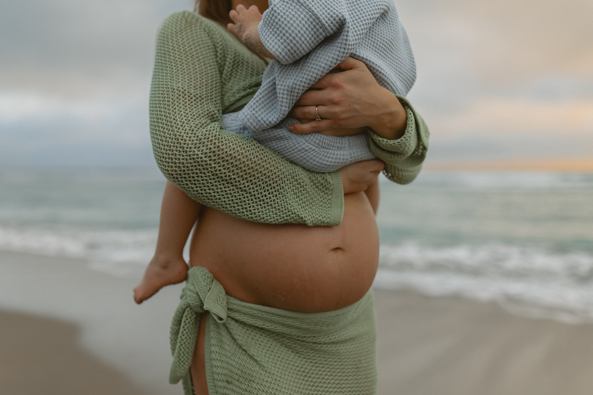 a detail shot of a pregnant woman wearing a two piece green crochet outfit holding her toddler above her belly