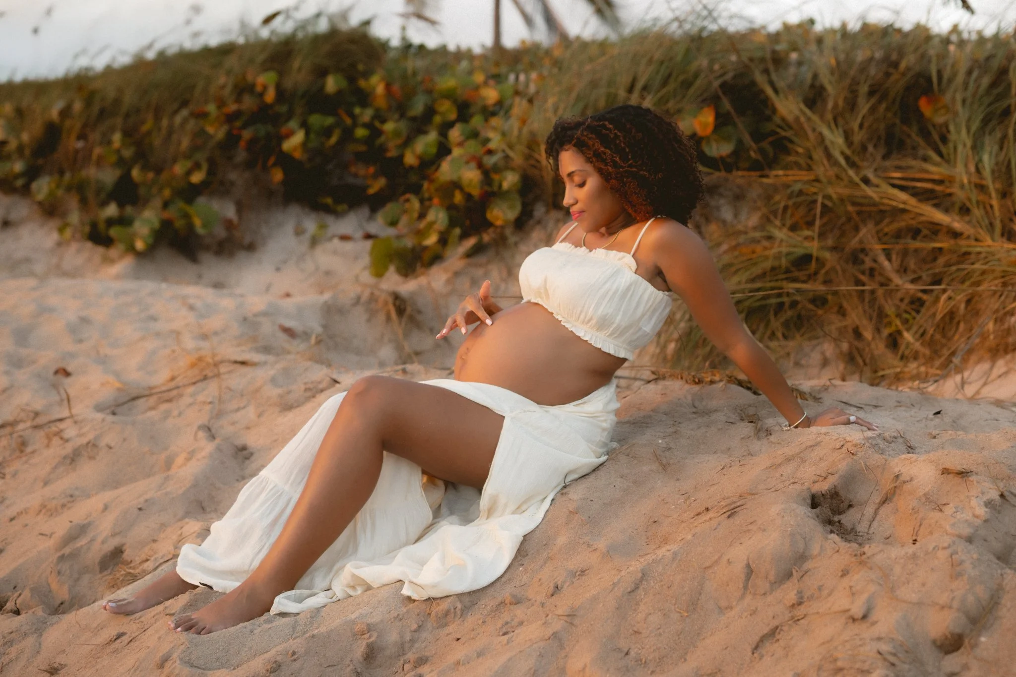 Pregnant woman sitting on sandy beach near sand dunes and coastal vegetation, wearing a white dress, looking at her belly.