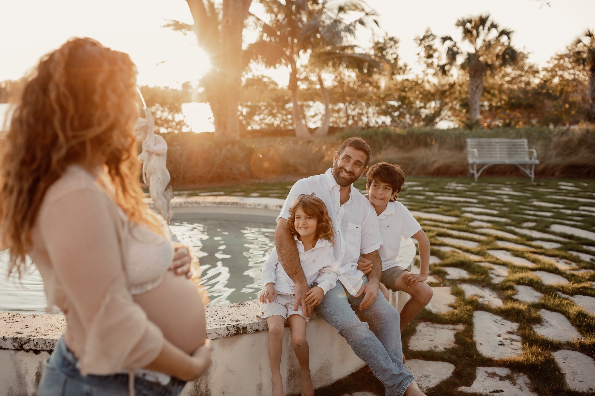 A pregnant woman with red hair standing in the foreground, facing a family of four sitting by a pond during sunset, with trees and a bench in the background.