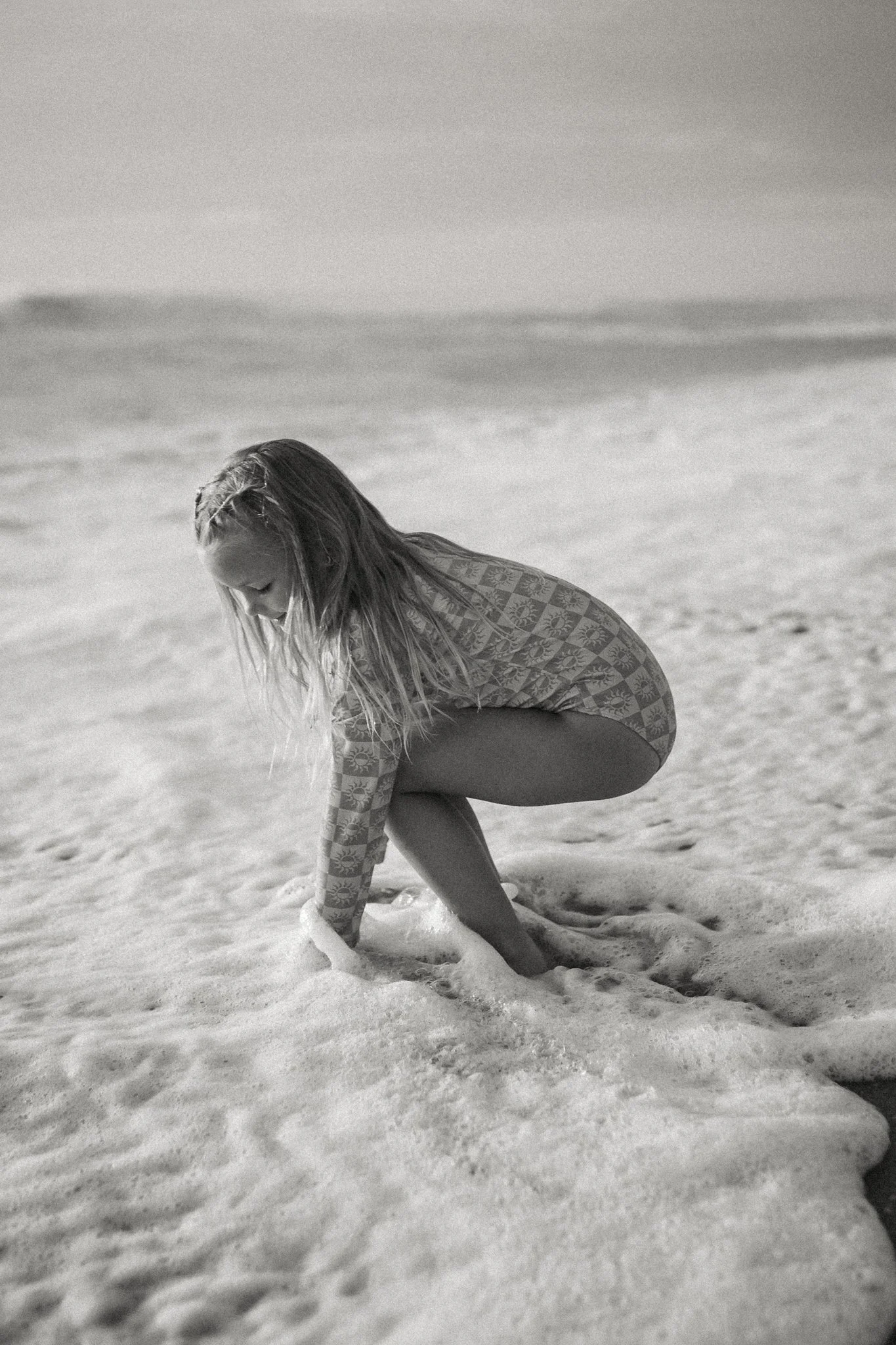 A young girl with long hair kneeling in the surf on the beach, wearing a long-sleeve patterned rash guard, and looking down at the water and foam around her legs.