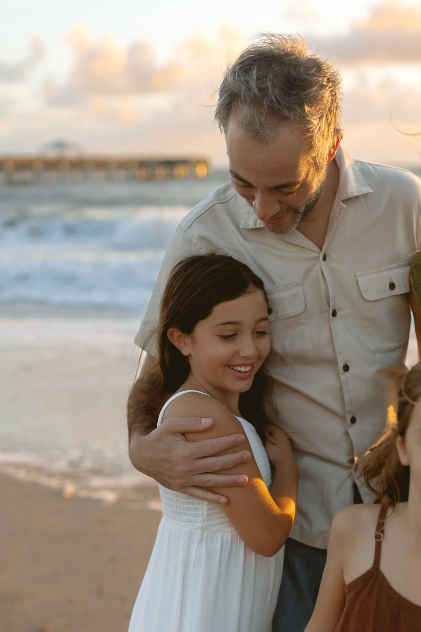 A man and a young girl are embracing on the beach at sunset, with the ocean and a pier in the background.