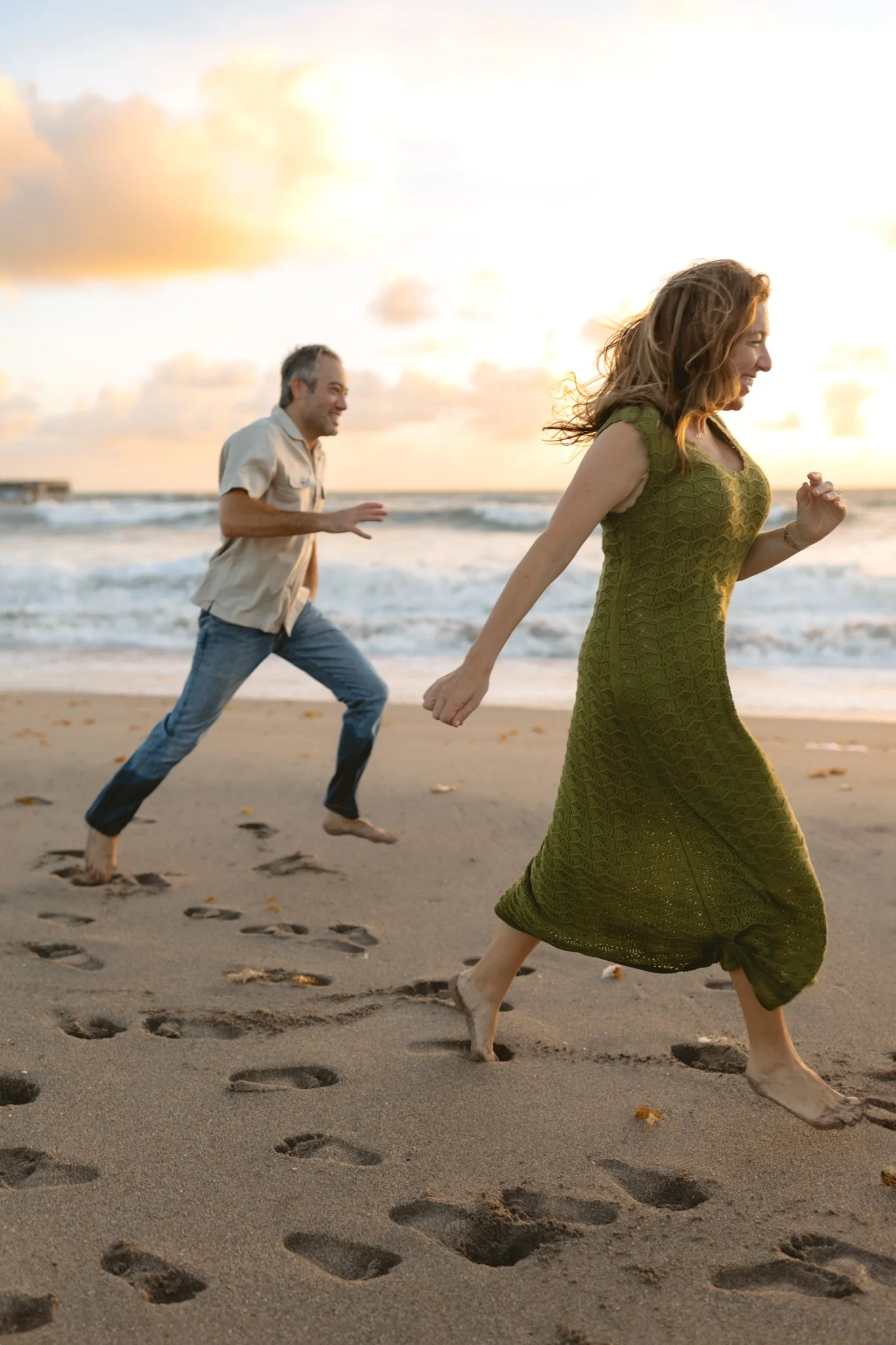 A woman and a man running barefoot on the beach at sunset, smiling and enjoying themselves.