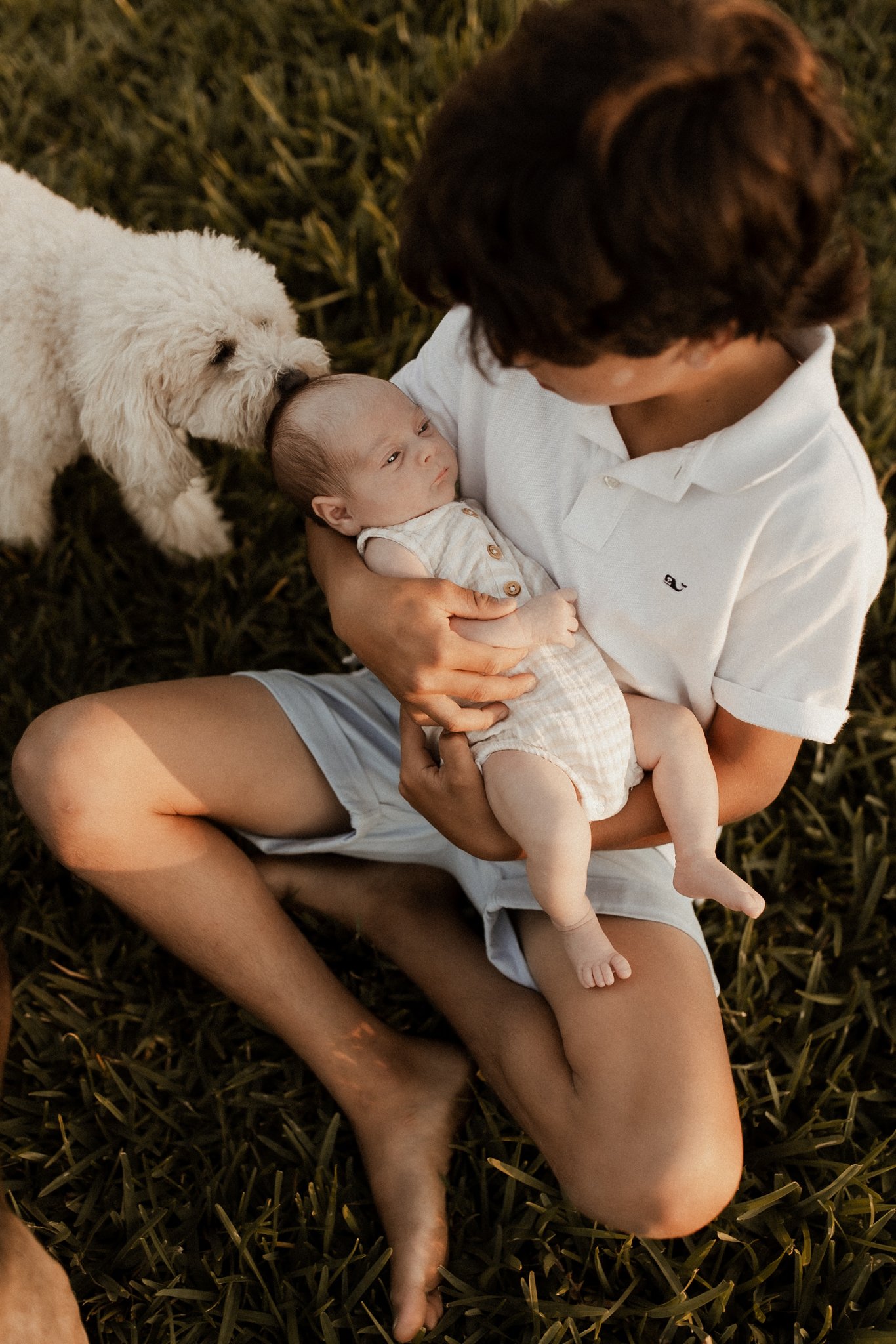 A young boy is holding a baby while sitting on grass, with a small white puppy nearby.