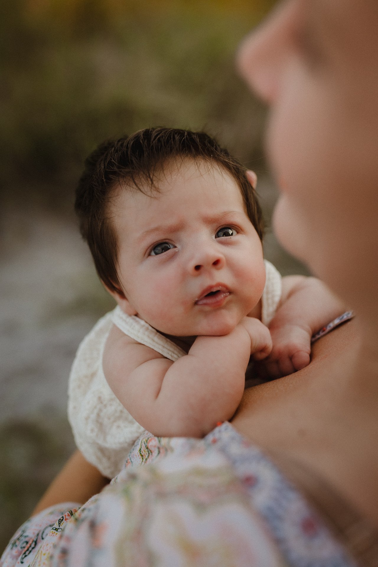 A close-up of a baby with dark hair looking up with a curious and slightly concerned expression, being held by an adult.
