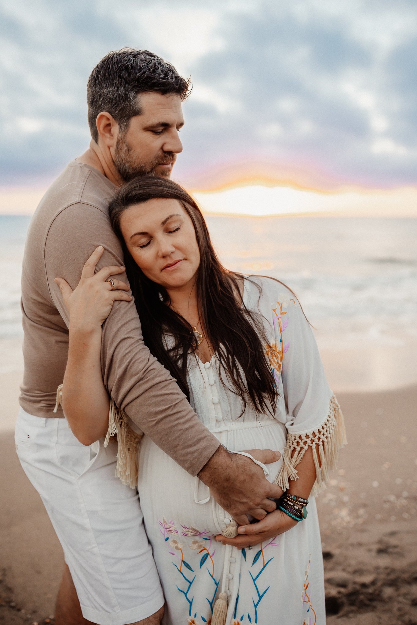 A man and pregnant woman embrace on the beach during sunset, with the ocean and sky in the background.