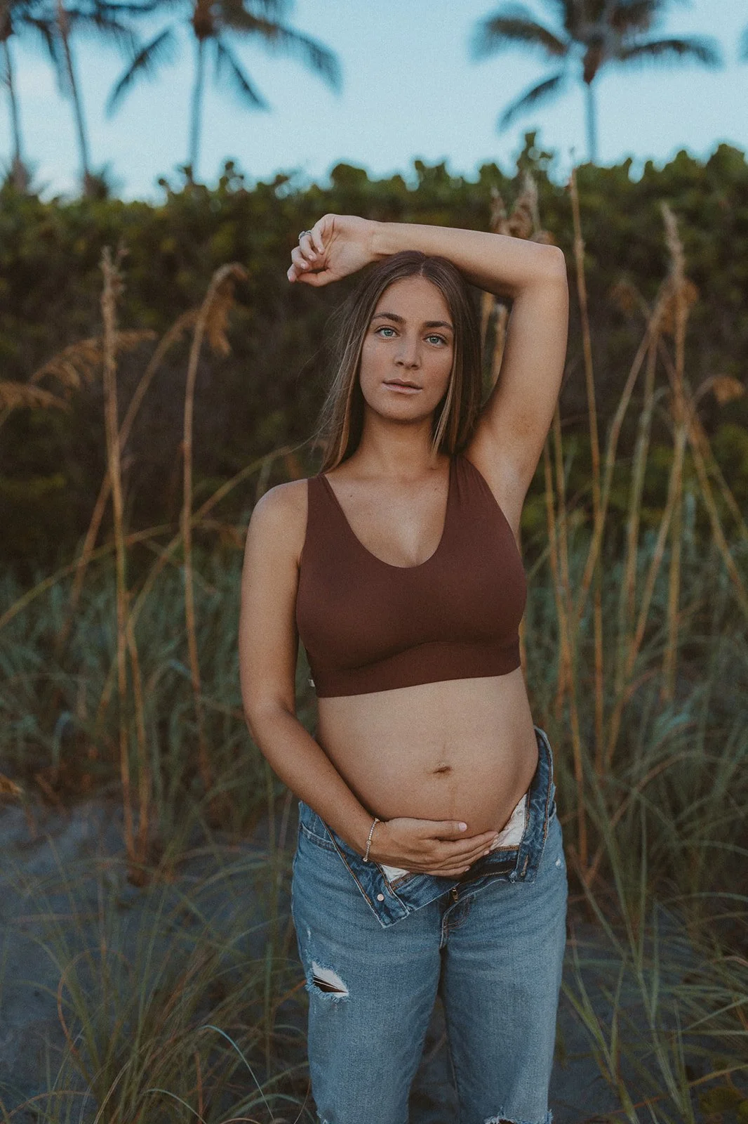 A pregnant woman standing outdoors in a field of tall grass and palm trees, holding her belly with one hand and raising her other arm above her head.