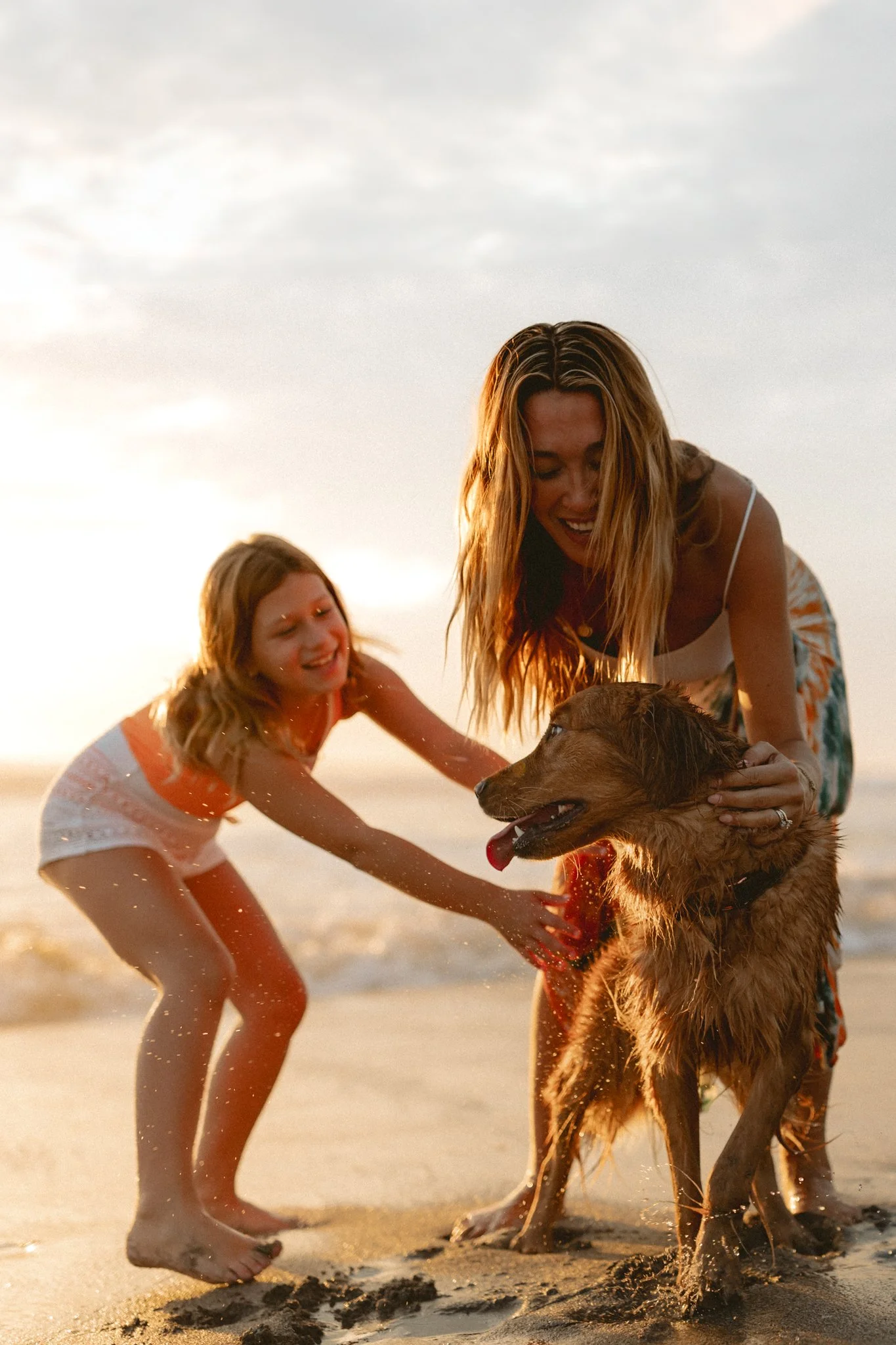A woman and a girl playing with a wet dog on the beach during sunset.