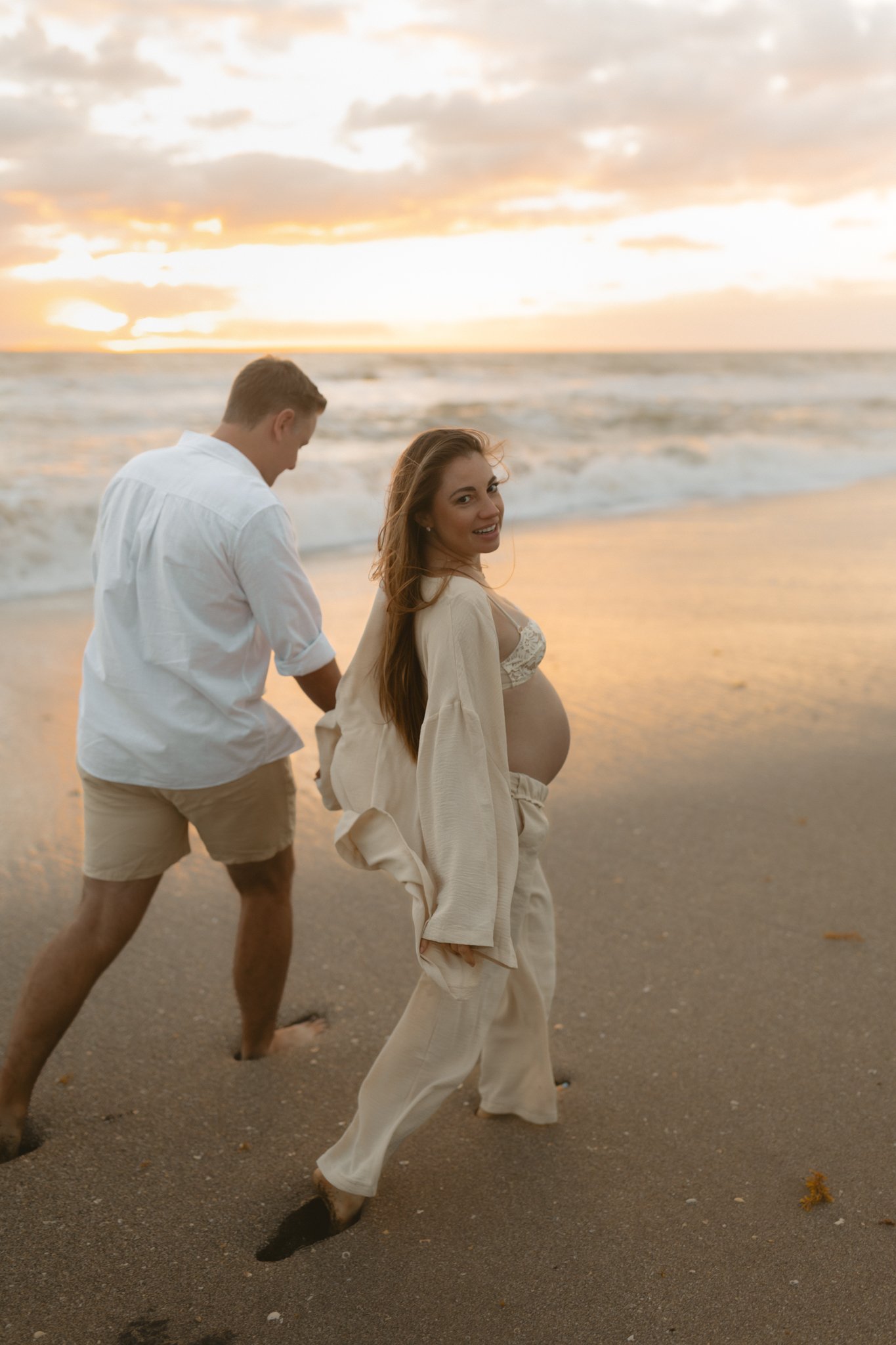 A pregnant woman and her partner on a beach at sunset, holding hands and walking in the sand.