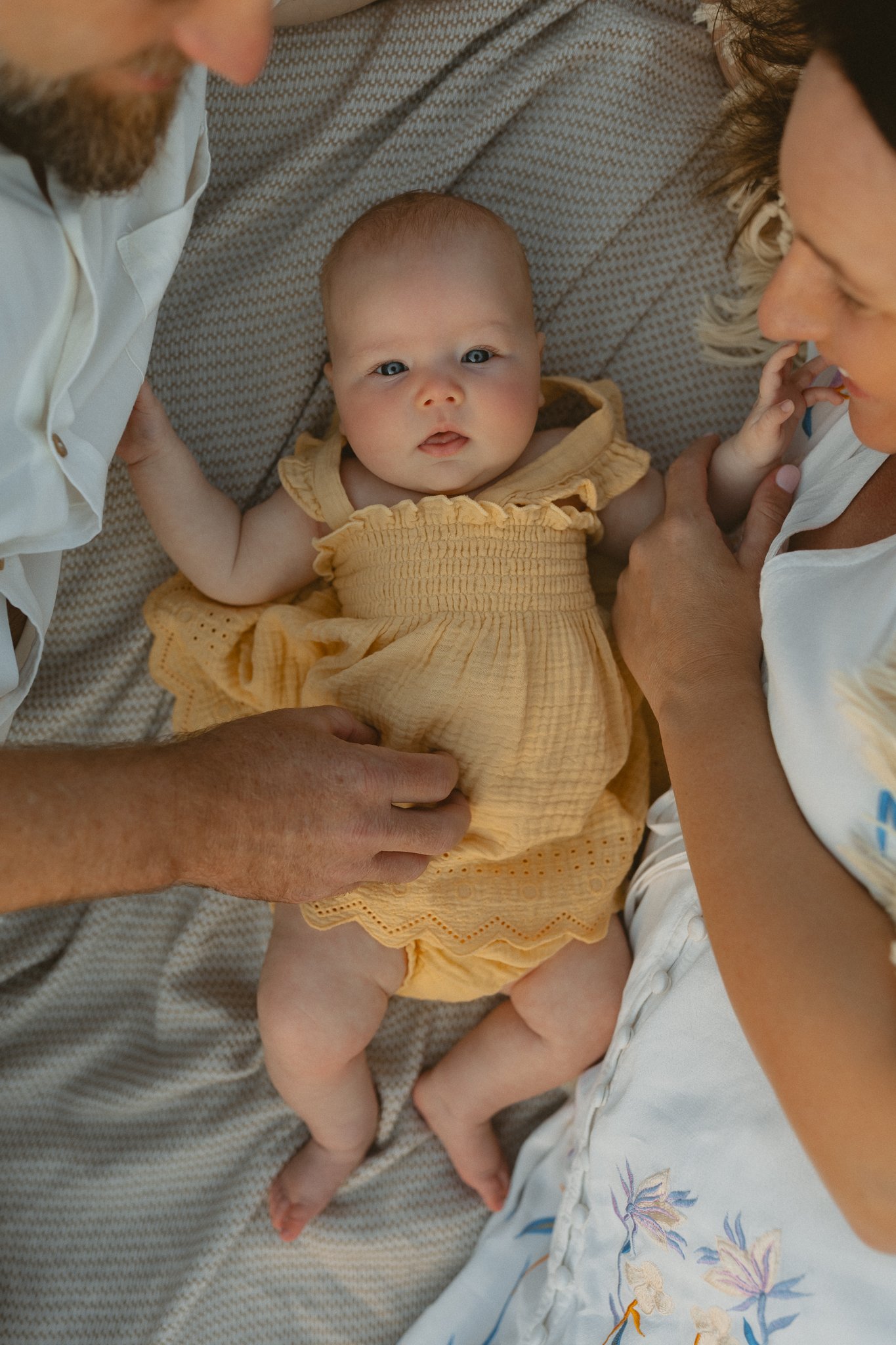 a baby dressed in a yellow outfit laying on a beach blanket in between their parents looking up at the camera