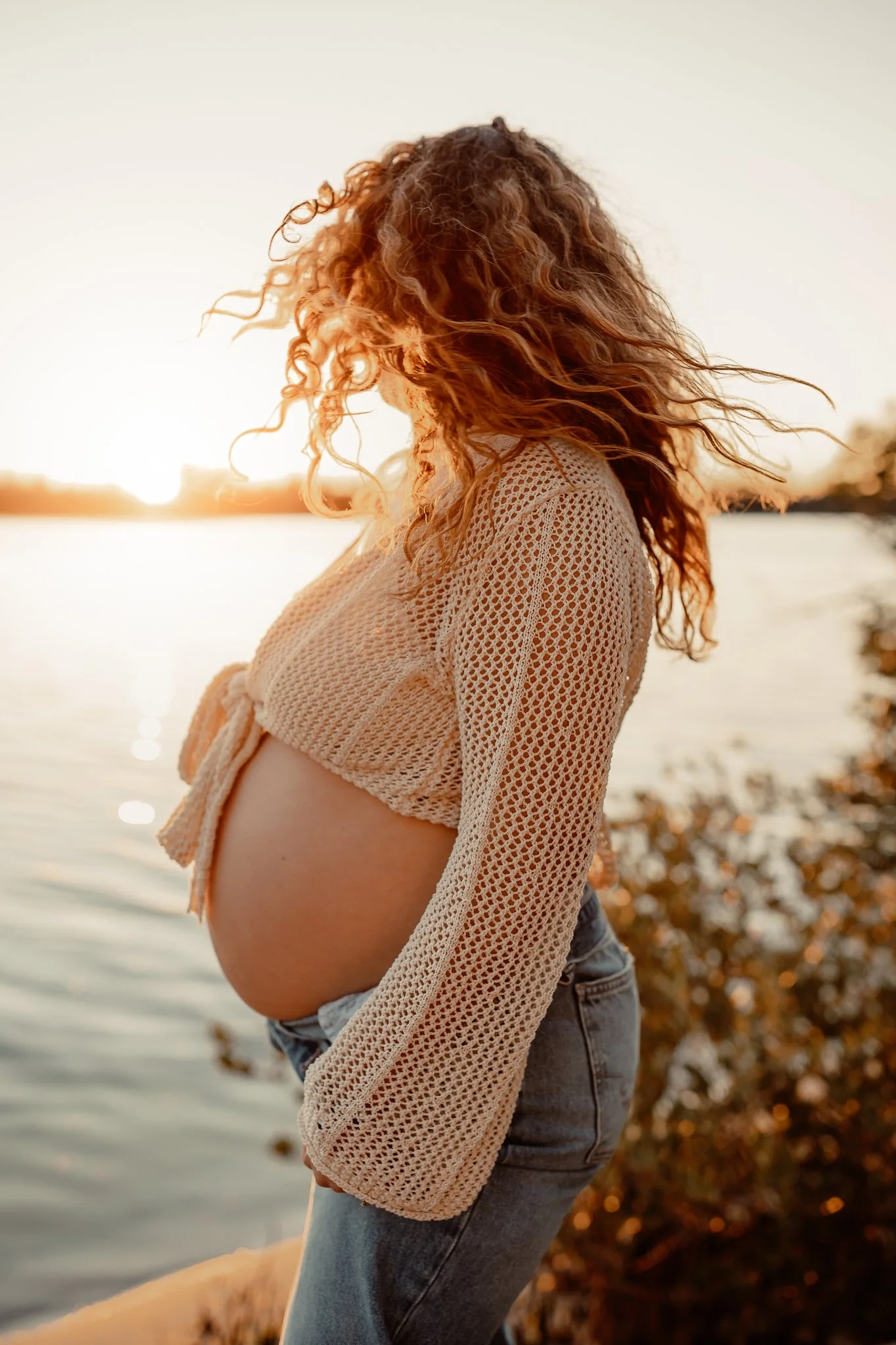 Pregnant woman with curly hair standing near a body of water during sunset