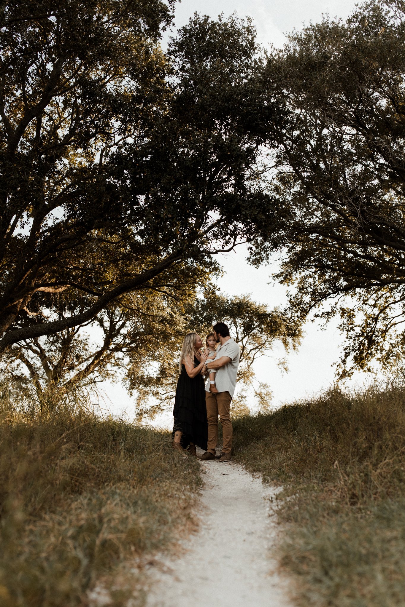 A family of three, a woman, a man, and a child, standing on a dirt path under large trees during sunset.