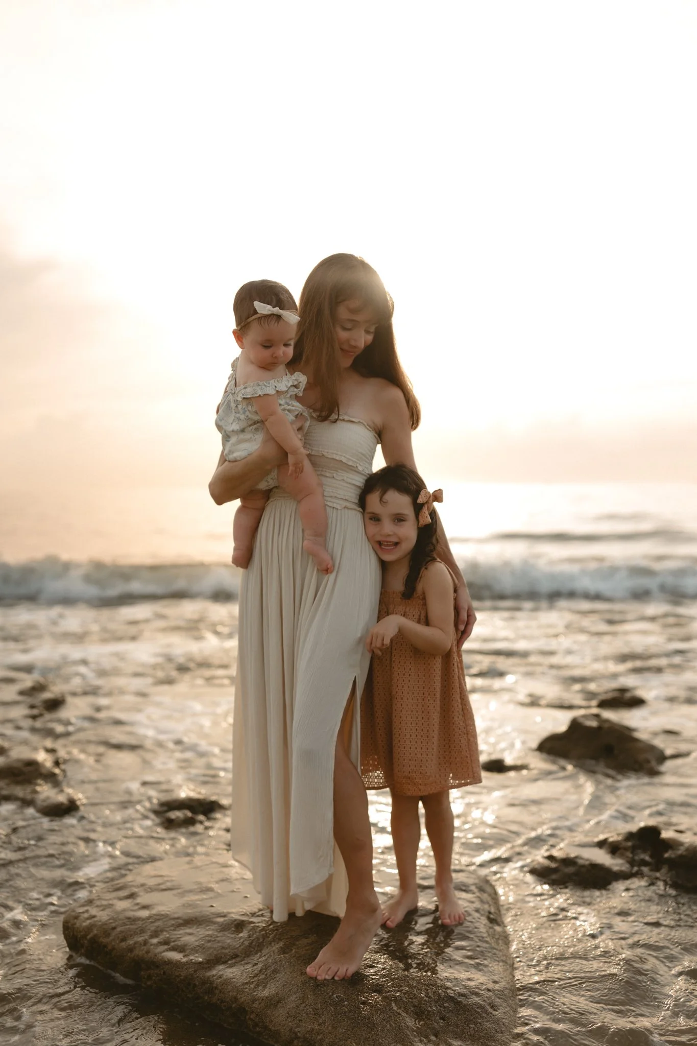 A woman with two young girls standing on a rock at the beach during sunset.