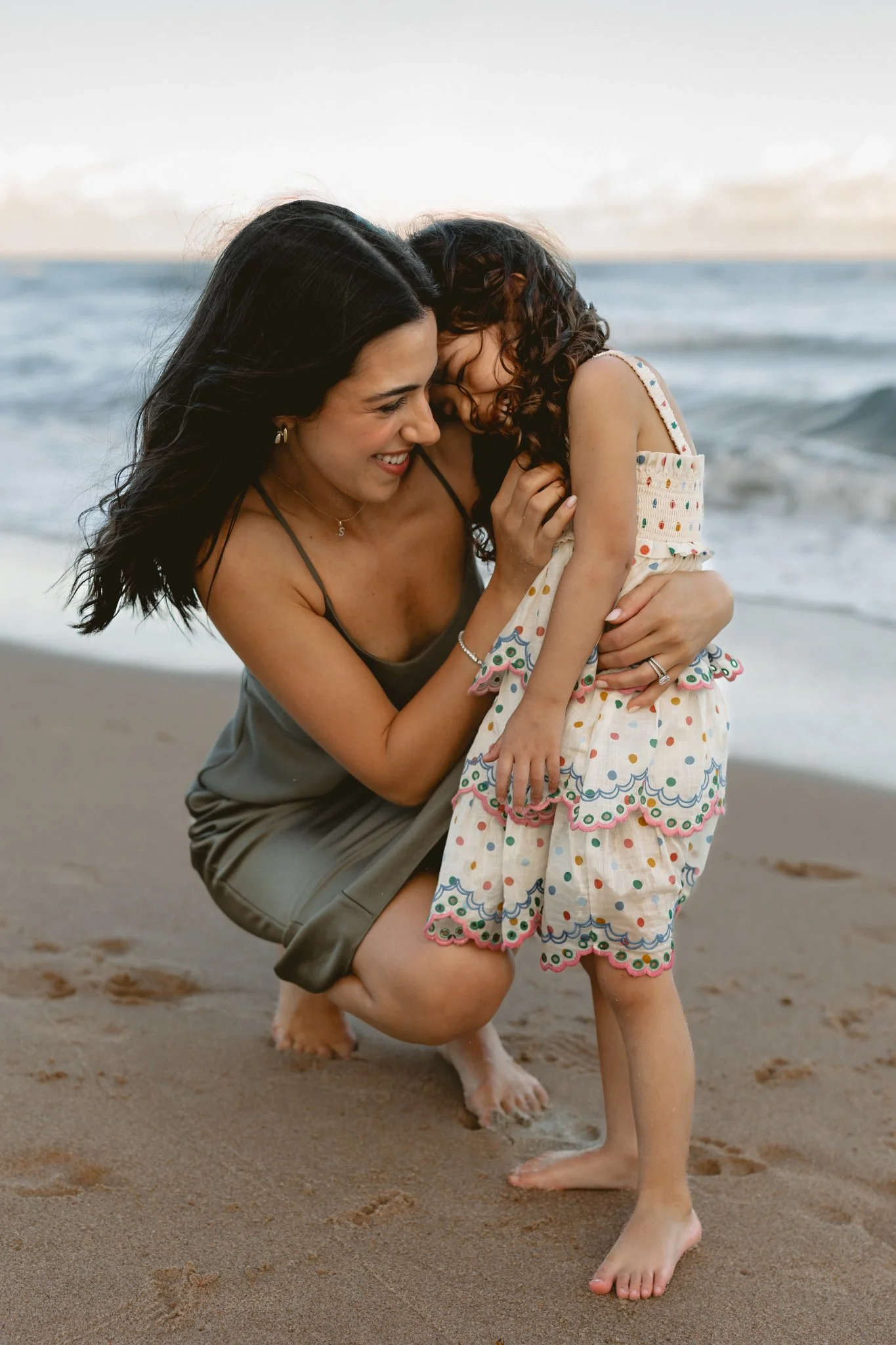 a mother kneeling down and giving her young daughter a hug on the beach during golden hour