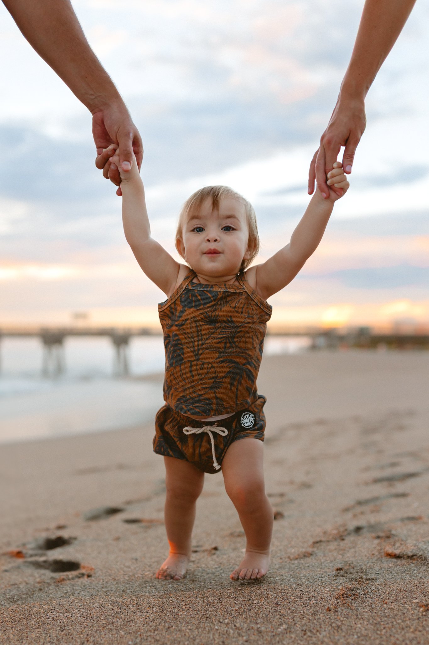 A toddler walking on the beach, holding hands with two adults, during sunset.