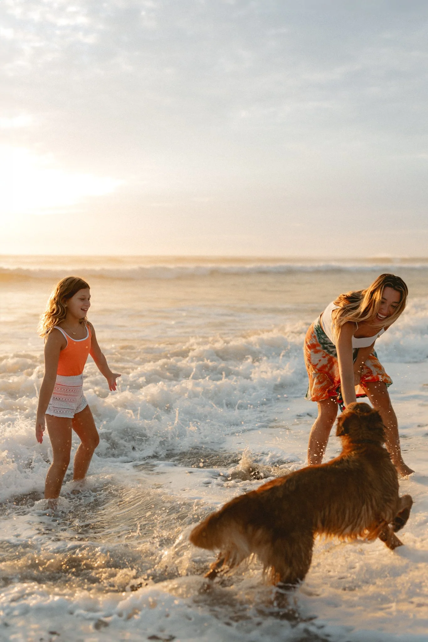 A woman and a young girl playing with a dog in the ocean at sunset, with waves splashing around them.