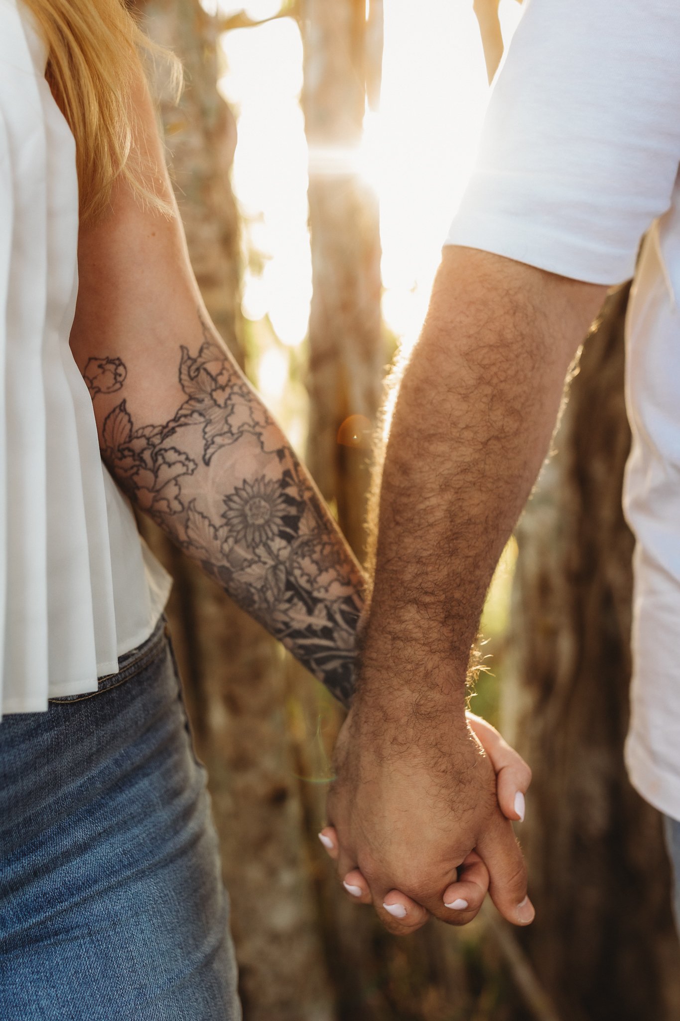 A couple holding hands outdoors, sunlight shining through trees, close-up of their intertwined hands and forearms.