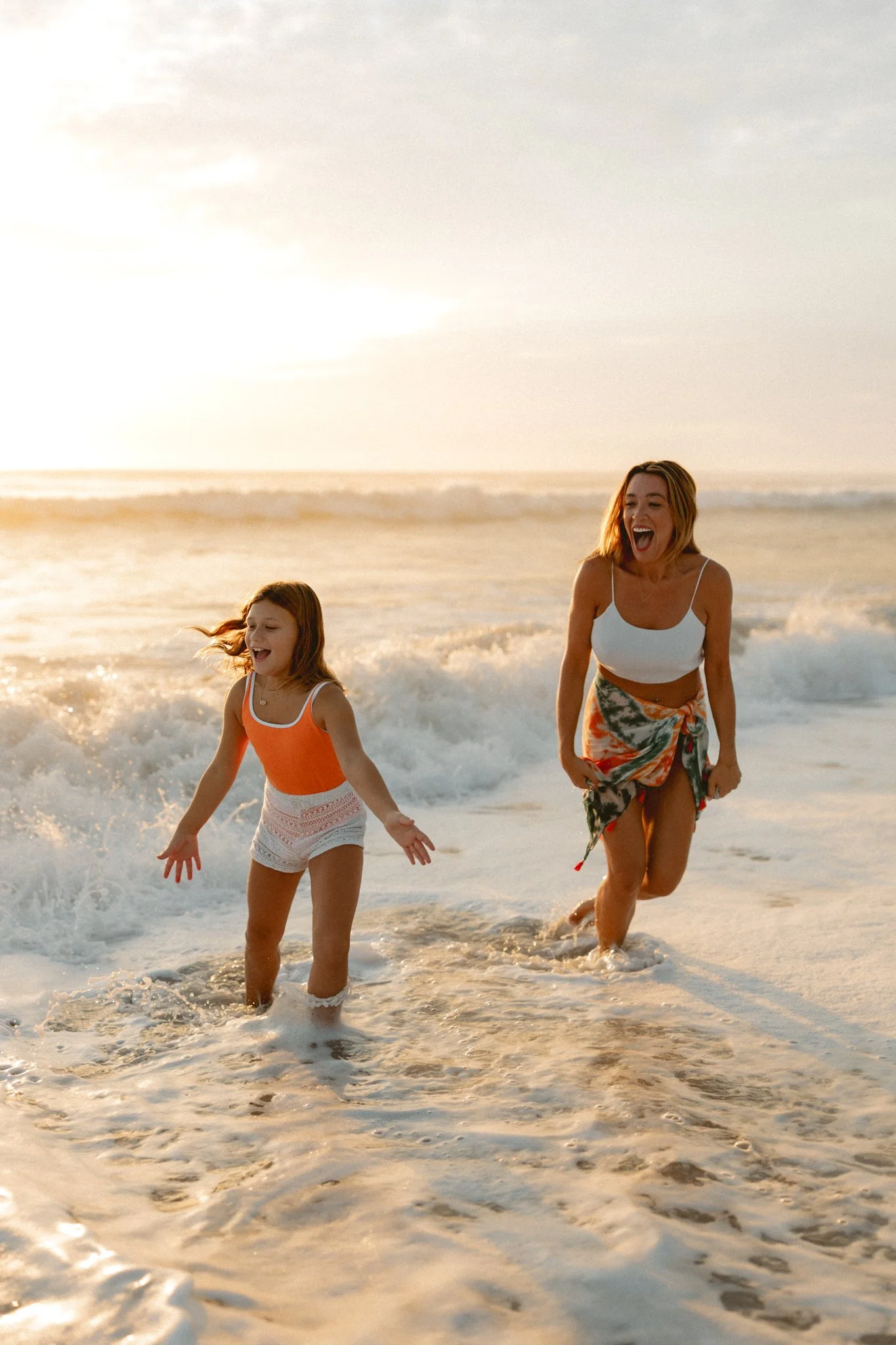 A woman and a young girl joyfully running through ocean waves at the beach during sunset.