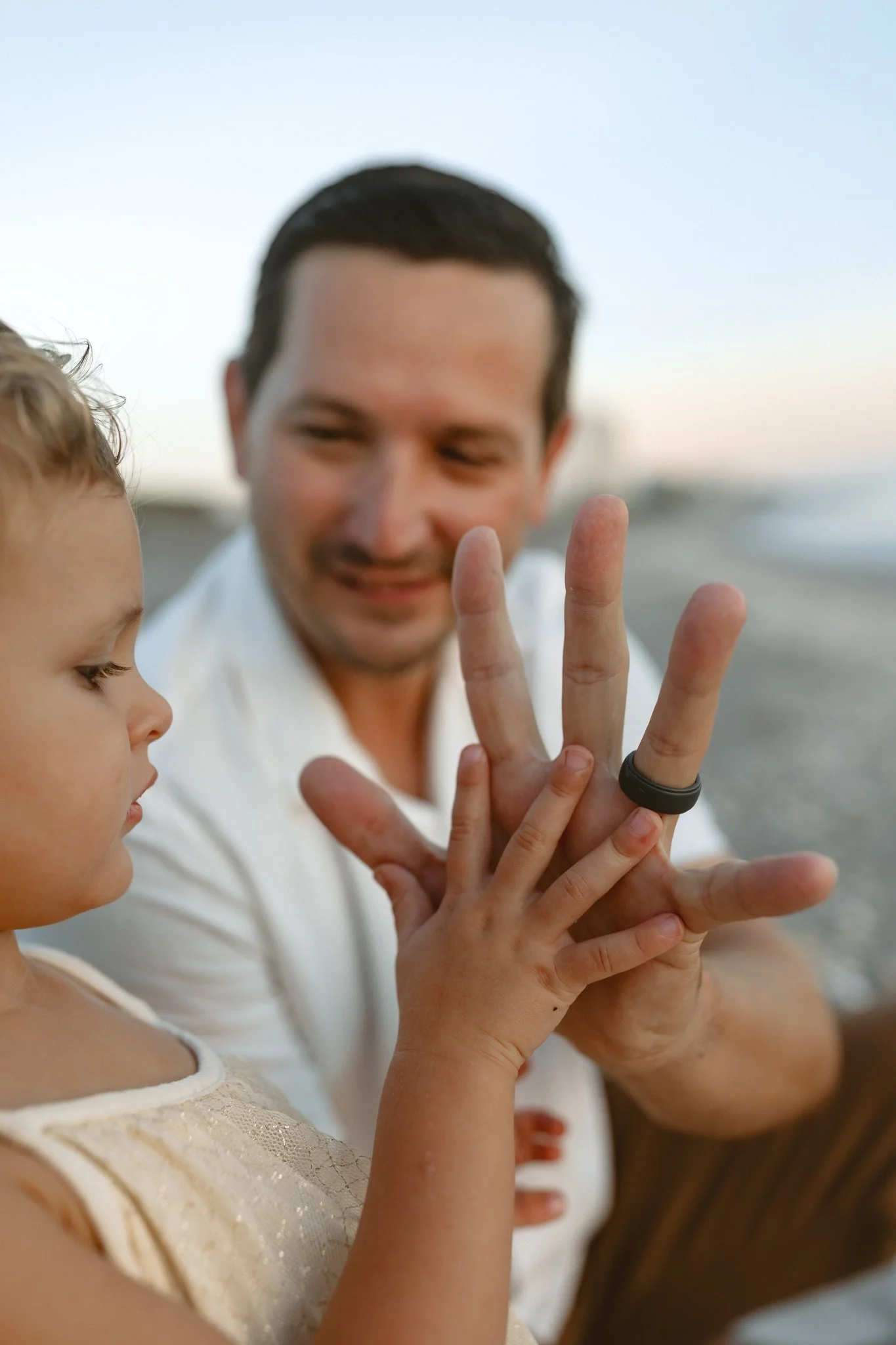 A man and a young girl are on a beach, playing with each other. The man is smiling and holding up his hand while the girl is touching his hand with her small fingers. The background shows the shoreline and the sky.