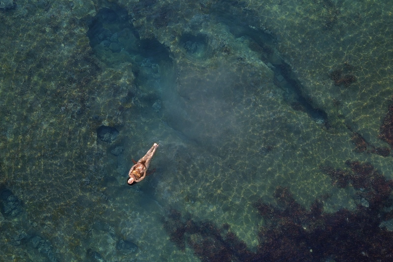 An aerial view of a woman snorkeling in clear, shallow water with rocks and seaweed visible beneath the surface.