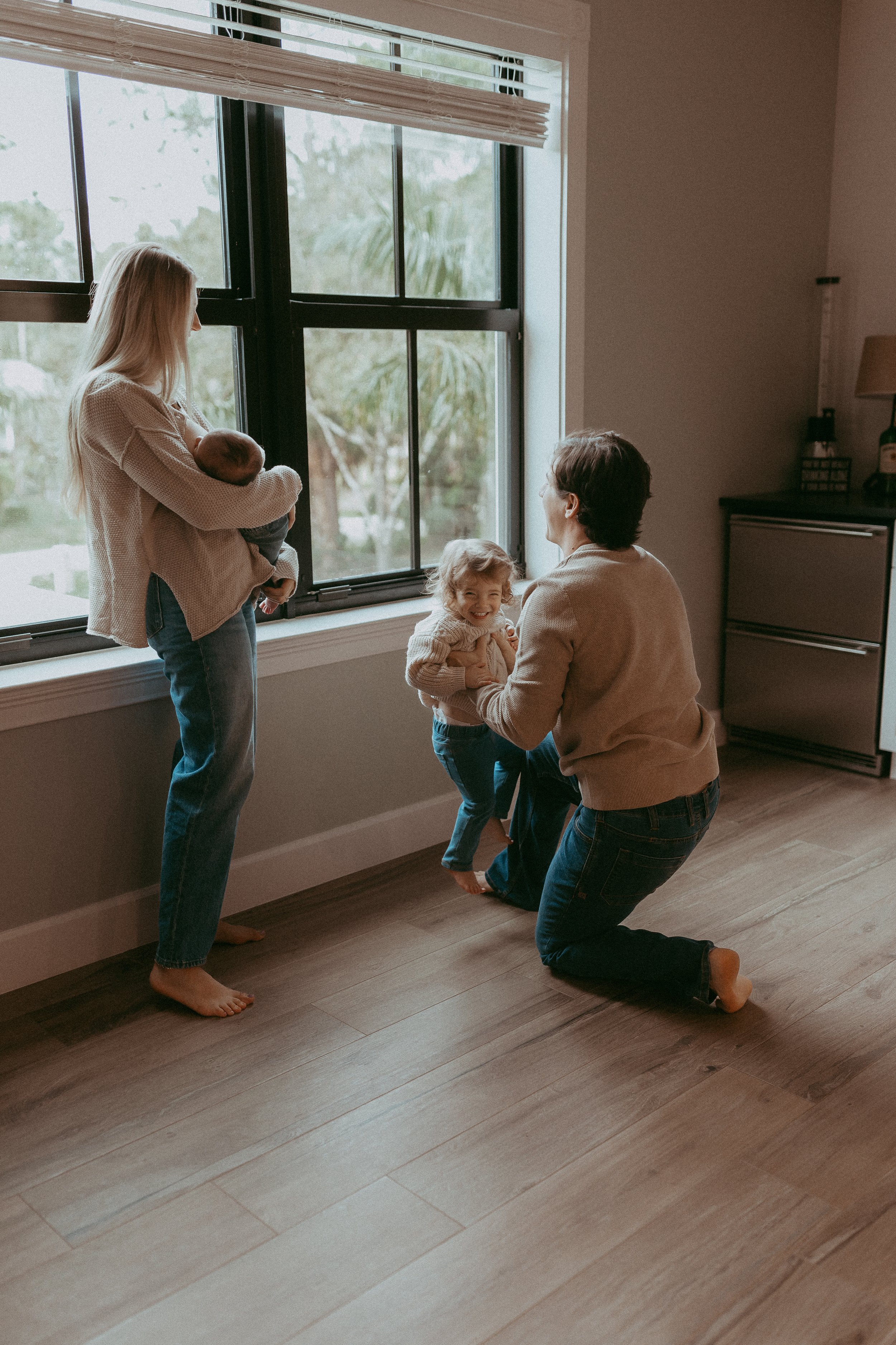A woman kneeling on the floor and a girl playing and smiling with a woman standing by the window holding a baby in her arms with another girl standing by her.