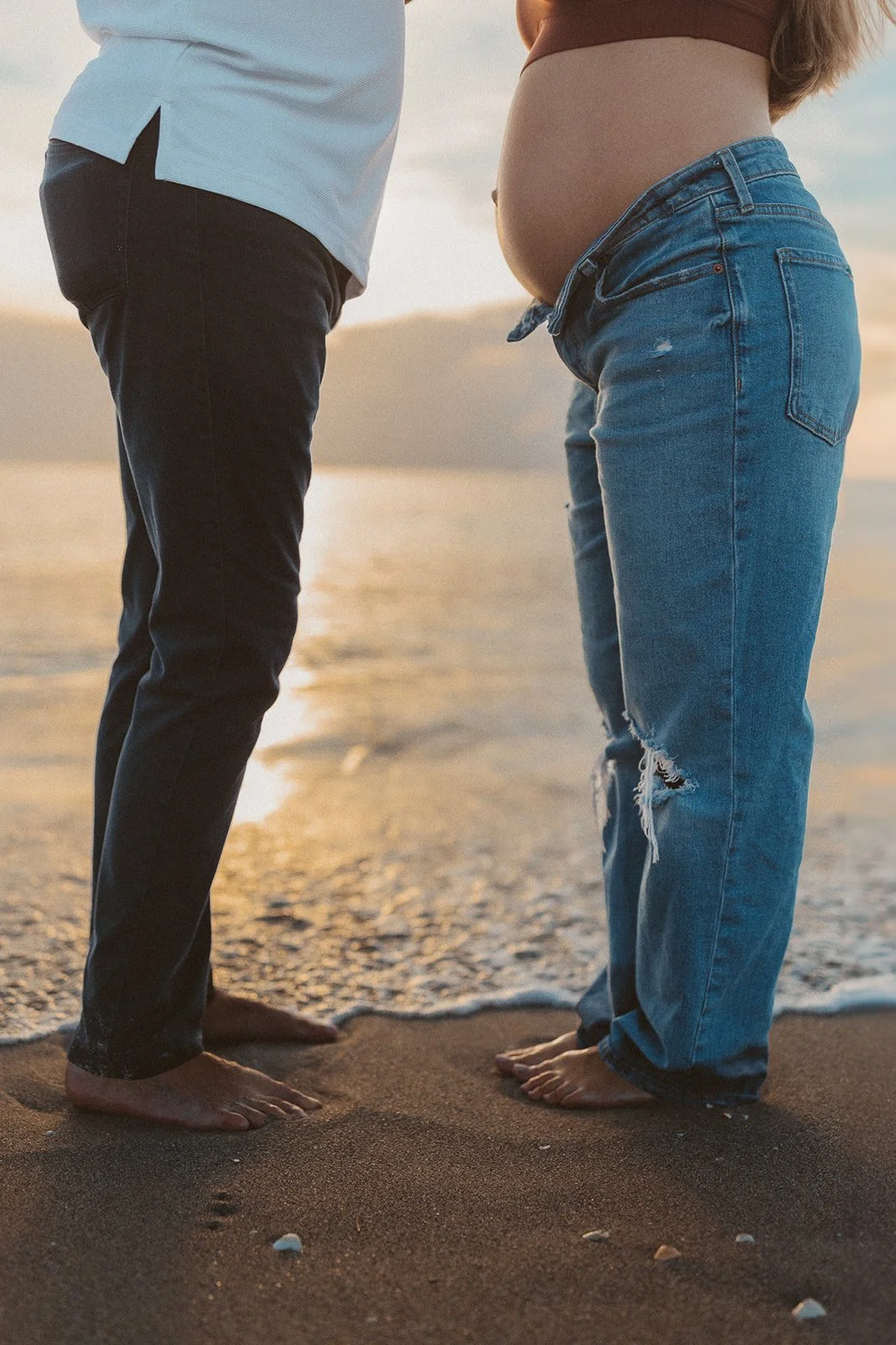 A pregnant woman and a man standing barefoot on the sand at the beach during sunset, facing each other.