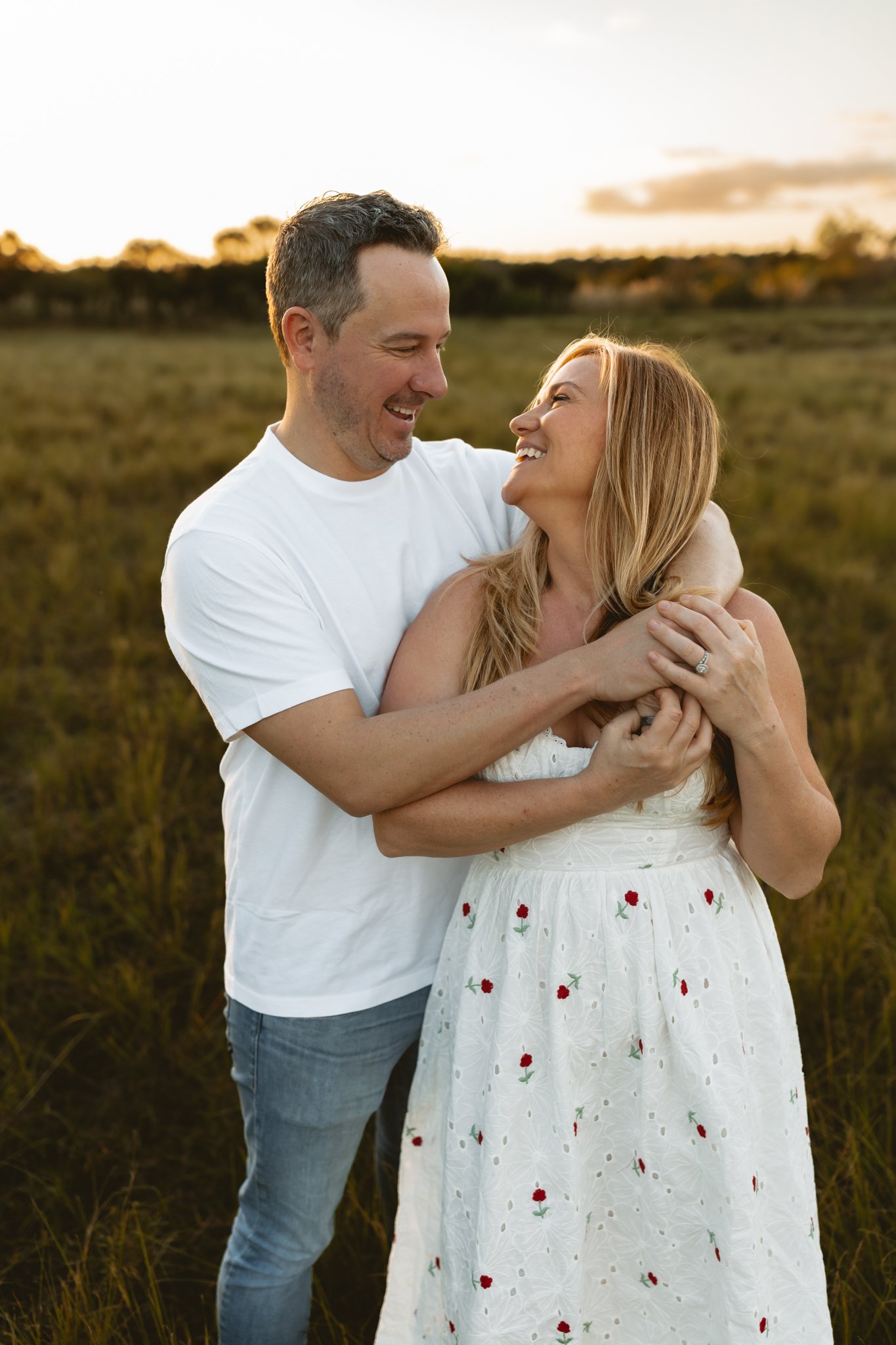 A couple standing in a field at sunset, smiling at each other, with the man holding the woman around her shoulders.