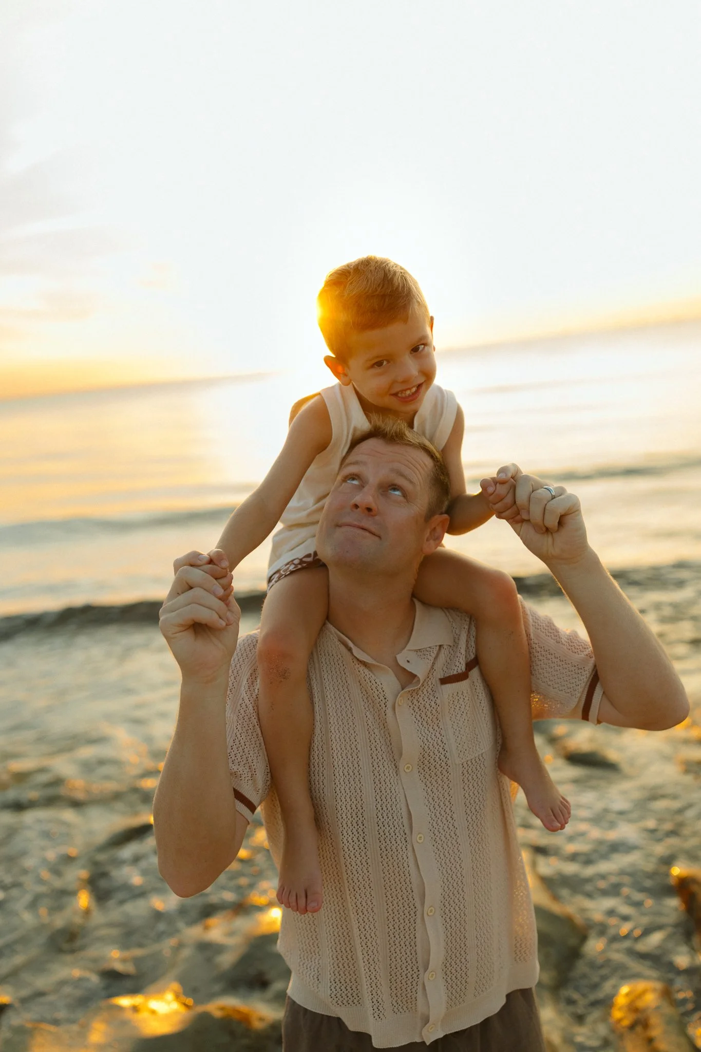 man with his young son on his shoulders and looking up to him while on the beach at sunrise