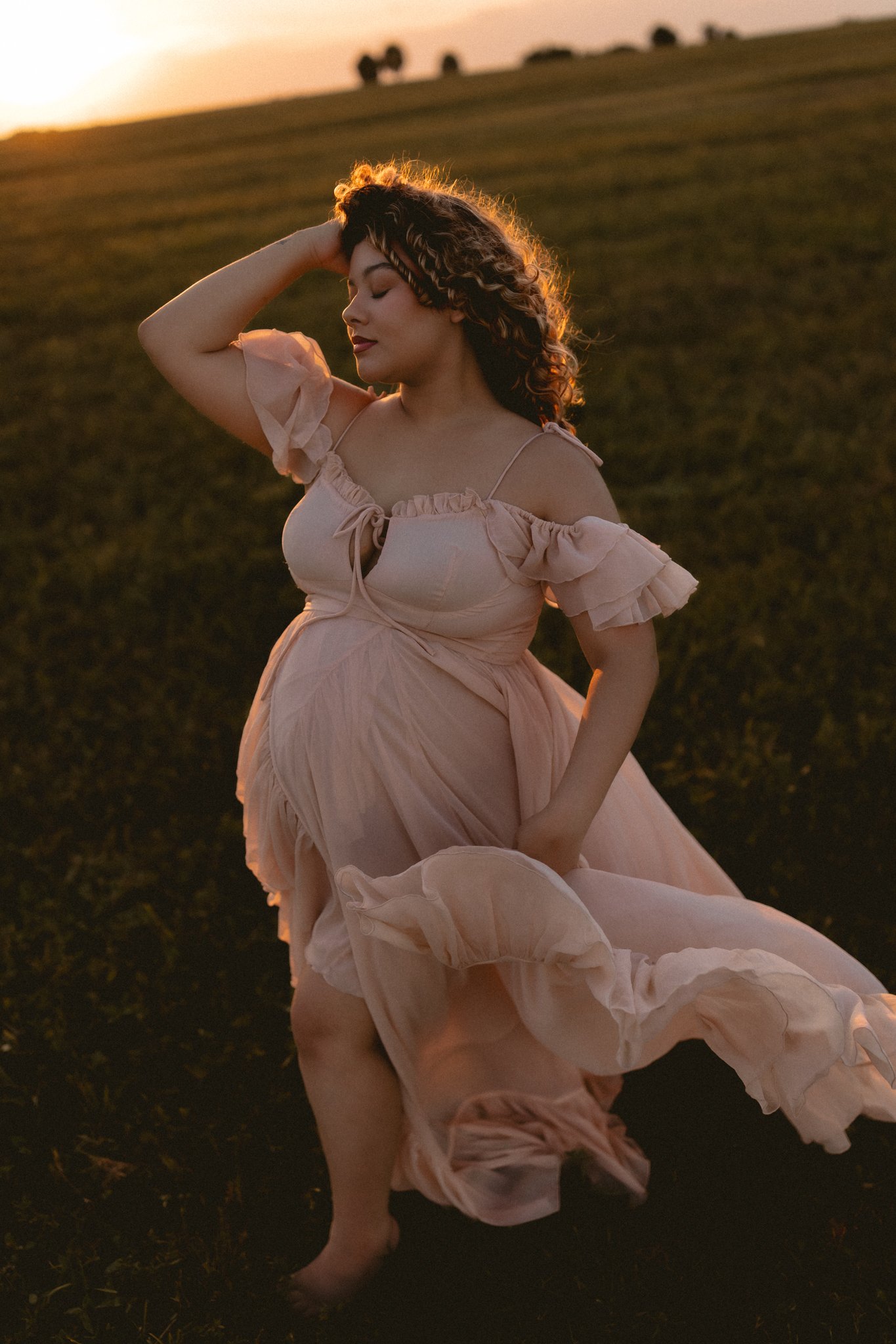 A woman in a flowing pink dress standing outdoors at sunset with her eyes closed and one hand resting on her head.