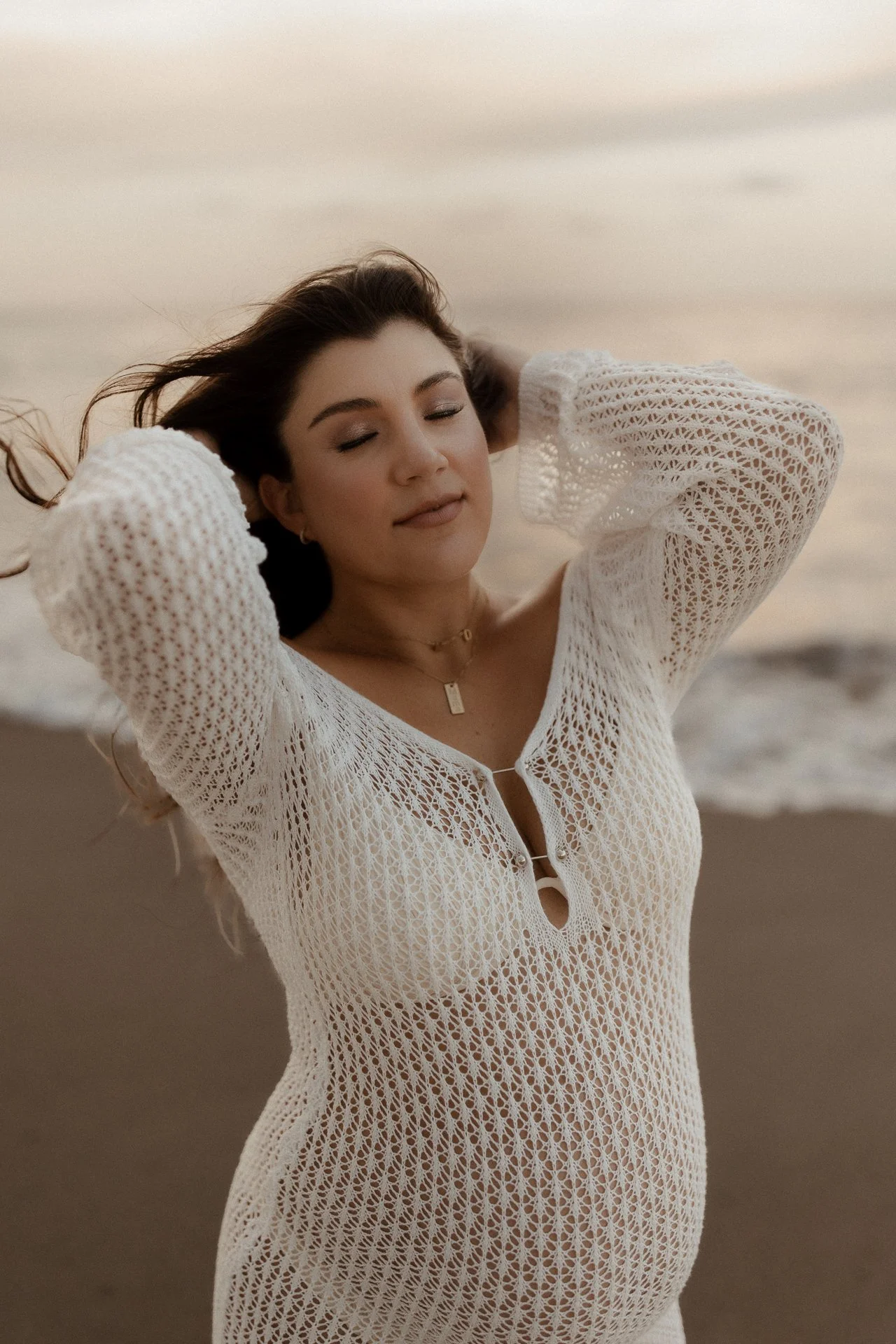 A woman with long brown hair relaxing on the beach during sunset, wearing a white, open-knit cover-up.