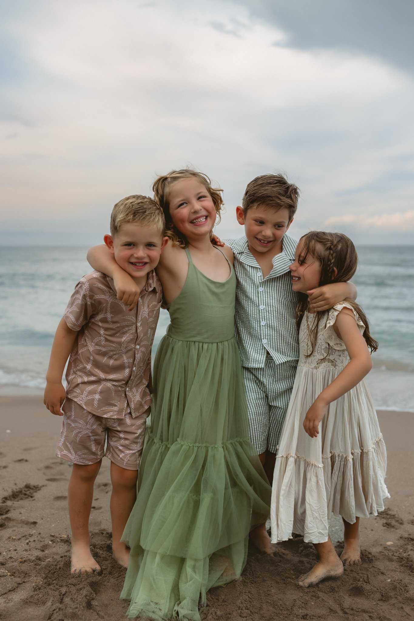 Four children hugging and smiling on the beach with ocean and cloudy sky in the background.