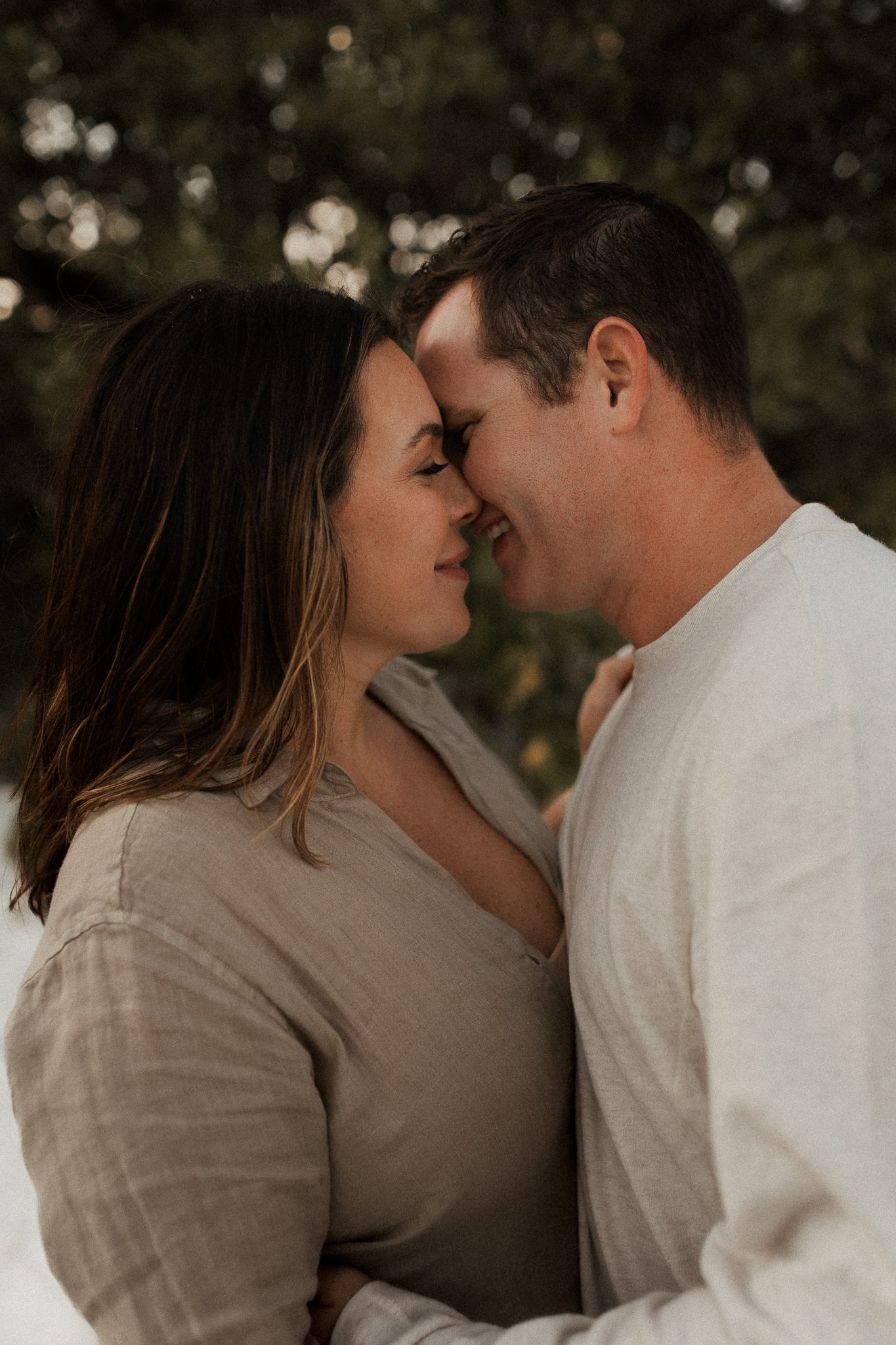A close-up of a couple with their foreheads touching and eyes closed, smiling lovingly outdoors during dusk.
