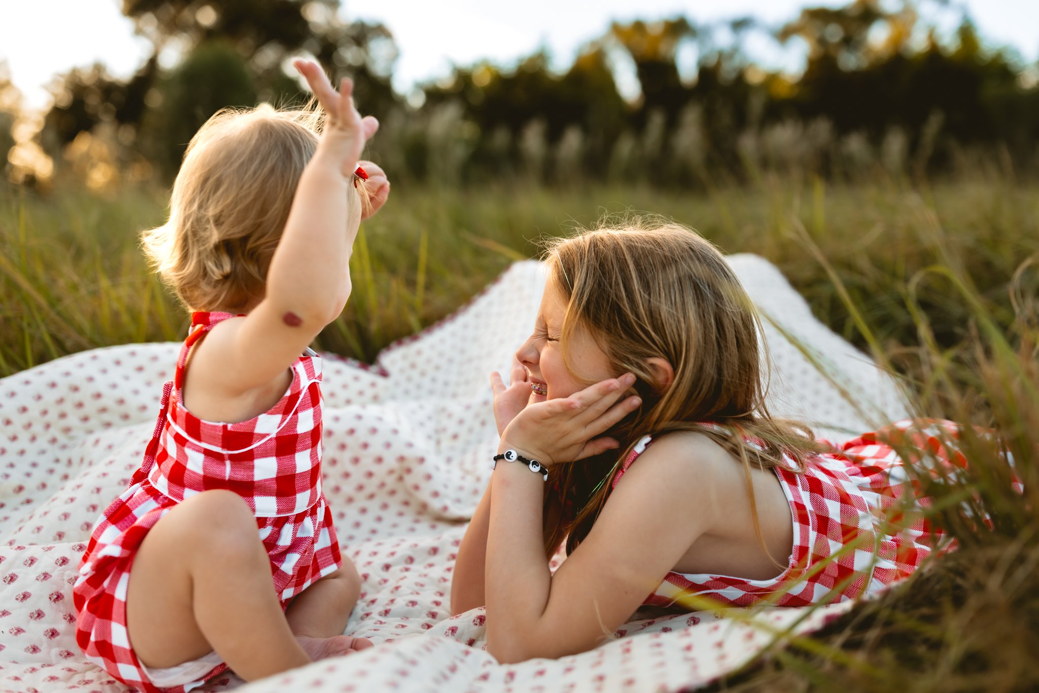 Two young girls in red and white checkered dresses lying on a blanket outdoors, playing and smiling at each other during sunset.