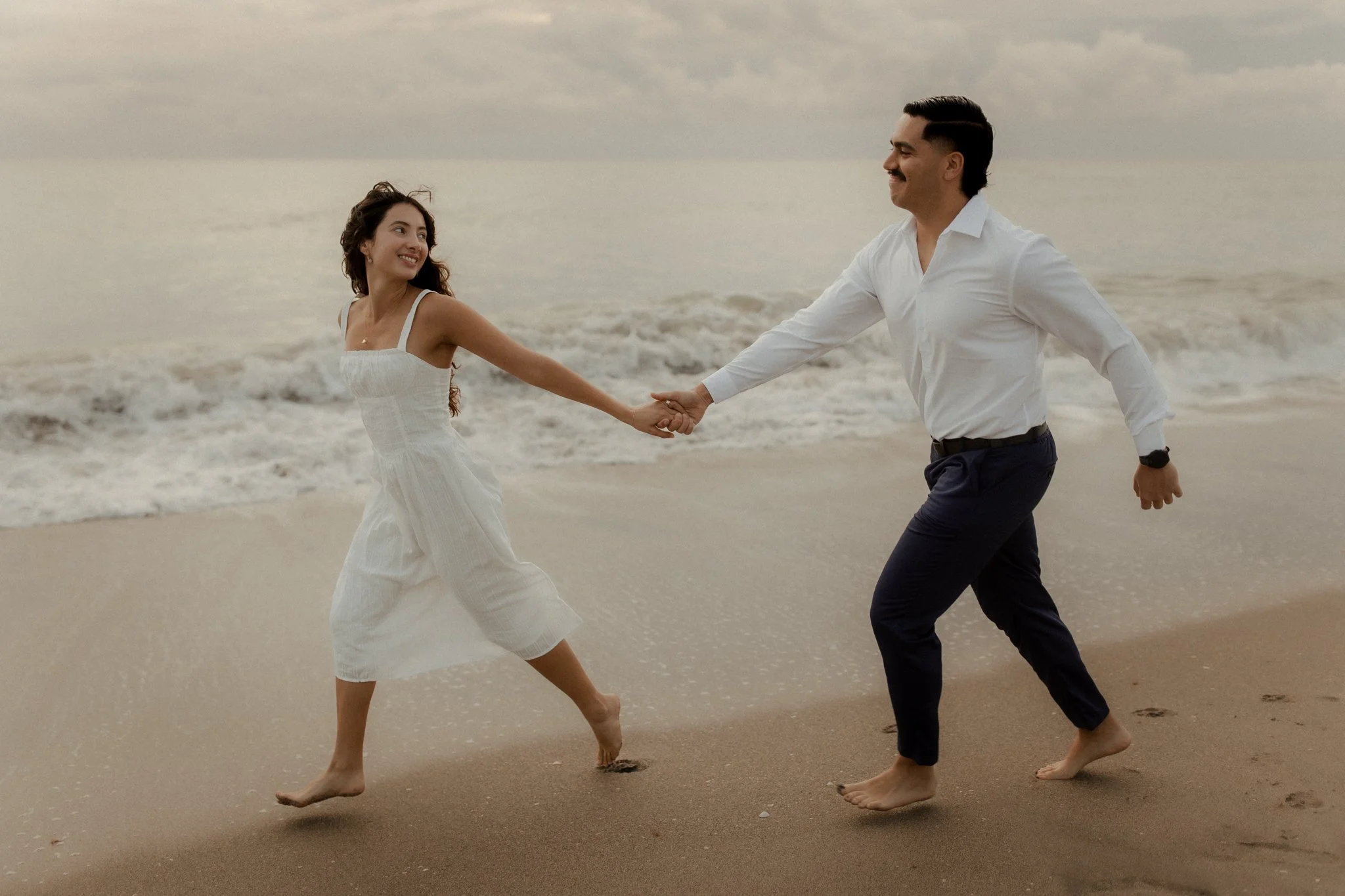 A couple is holding hands and running along the beach, with sand and ocean waves in the background, under a cloudy sky.