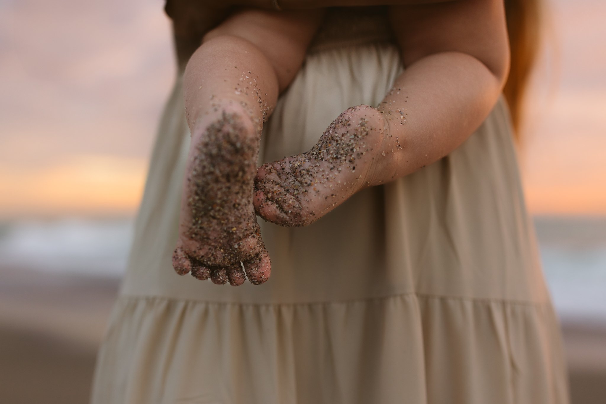 A close-up of a person's hand and fingers covered in sand, with a beach and sunset in the background.