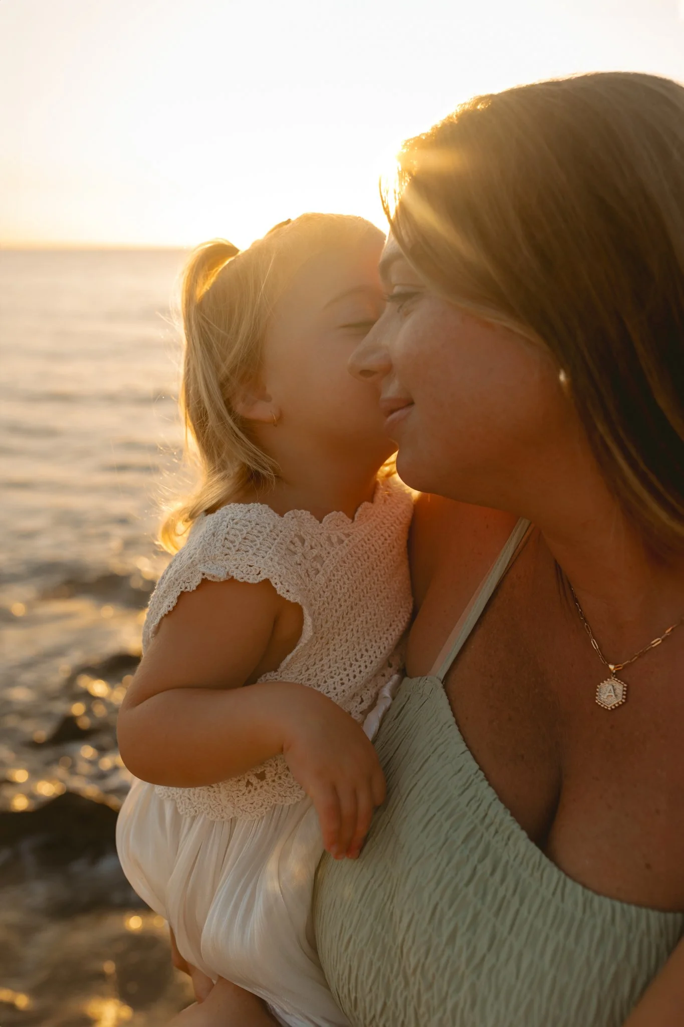 A woman holding a young girl on her shoulder at the beach during sunset, with them close to each other, eyes closed, sharing a tender moment.