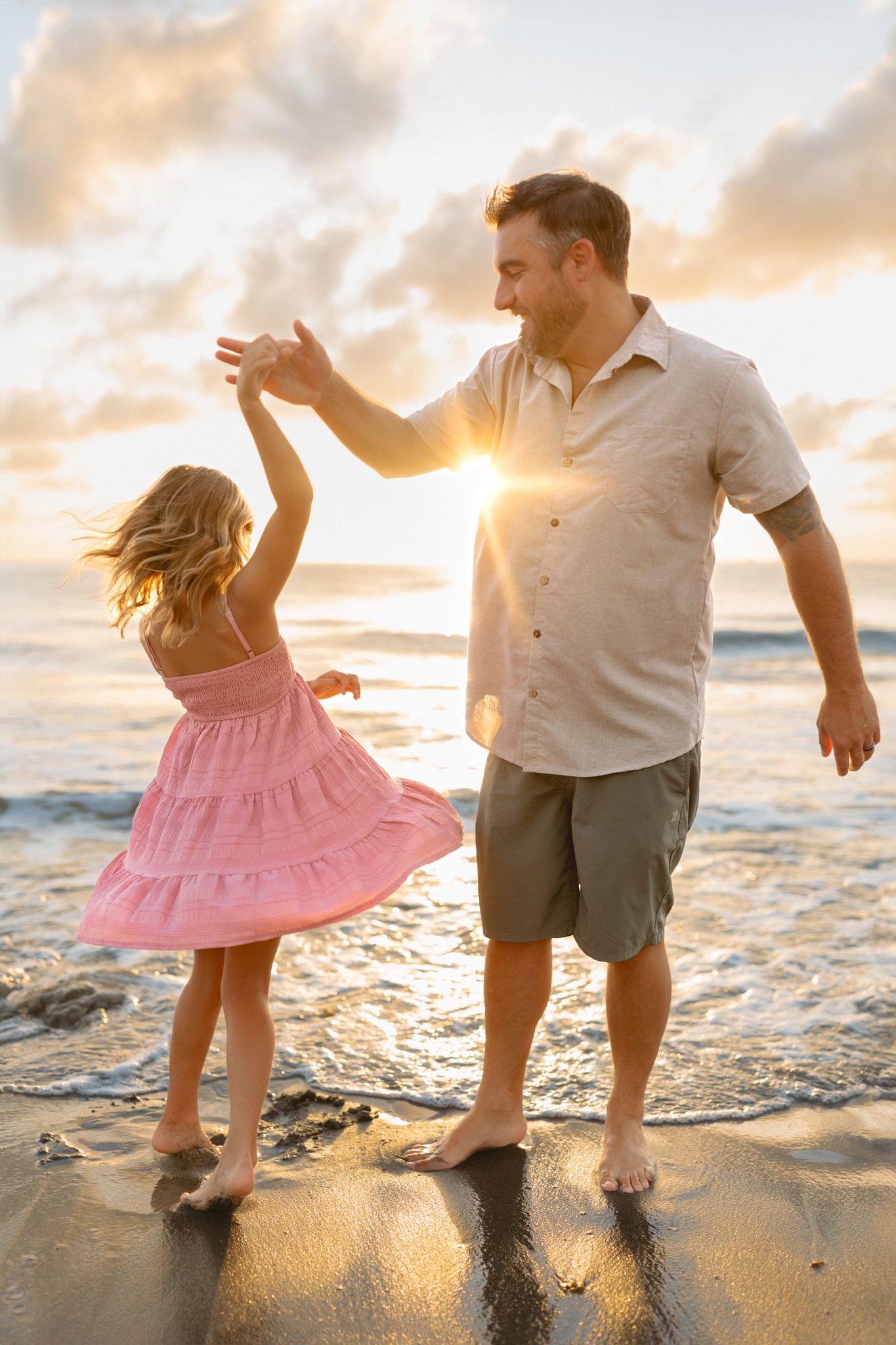 A man and a young girl dancing on the beach during sunset, holding hands, with the ocean in the background.