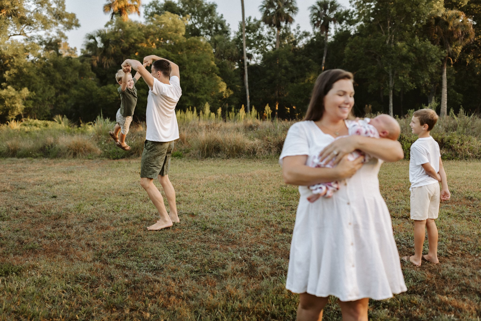 A family outdoor scene in the late afternoon with a woman holding a baby, a man twirling a young girl, and a boy standing nearby in a grassy field with trees in the background.