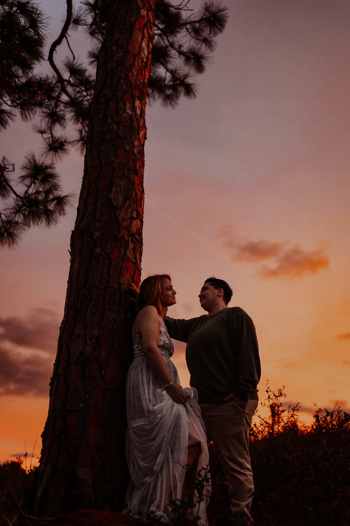 A man and woman stand close together outdoors at sunset, leaning against a large tree, looking at each other and smiling.